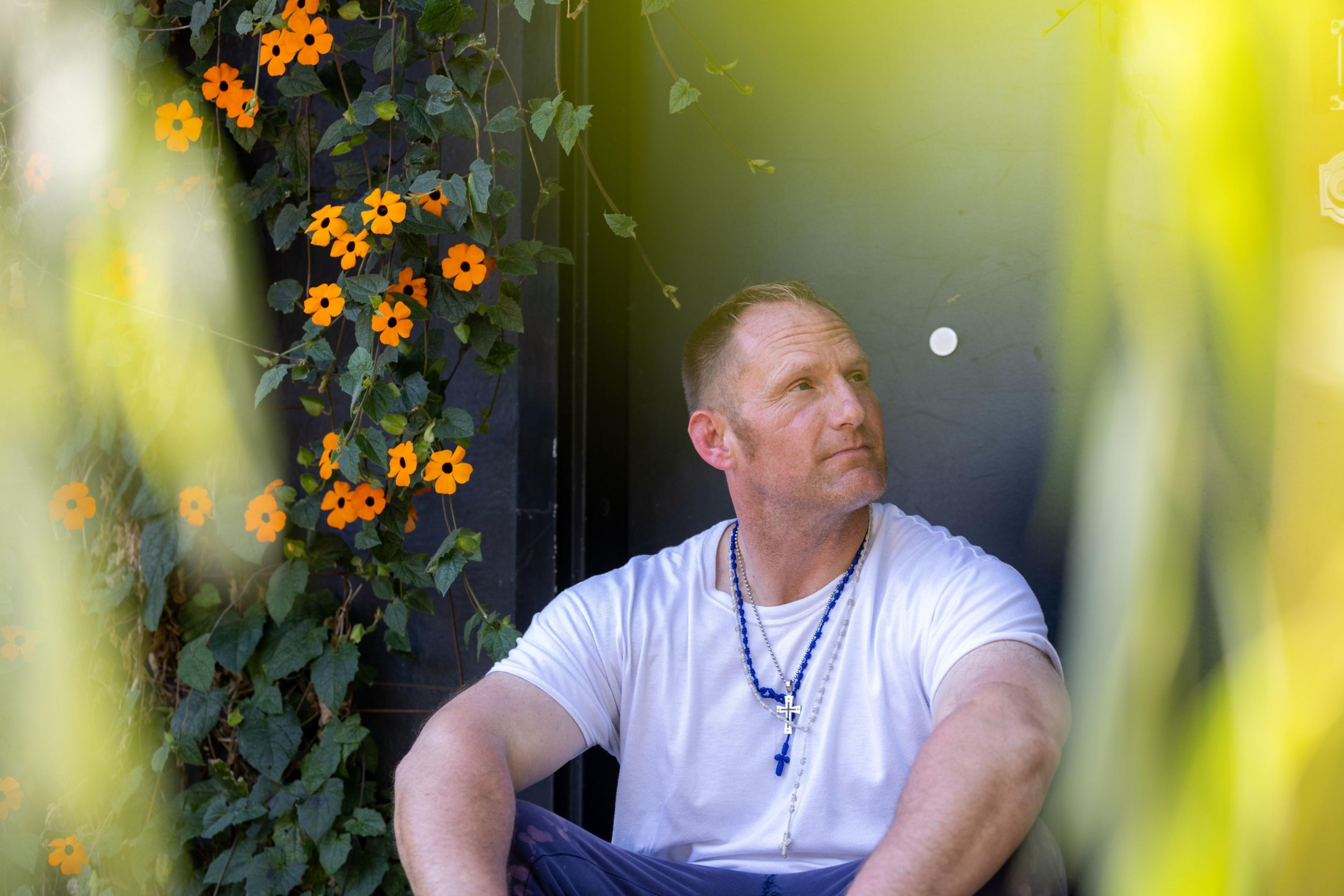 A man sits in a garden of flowers and greenery.