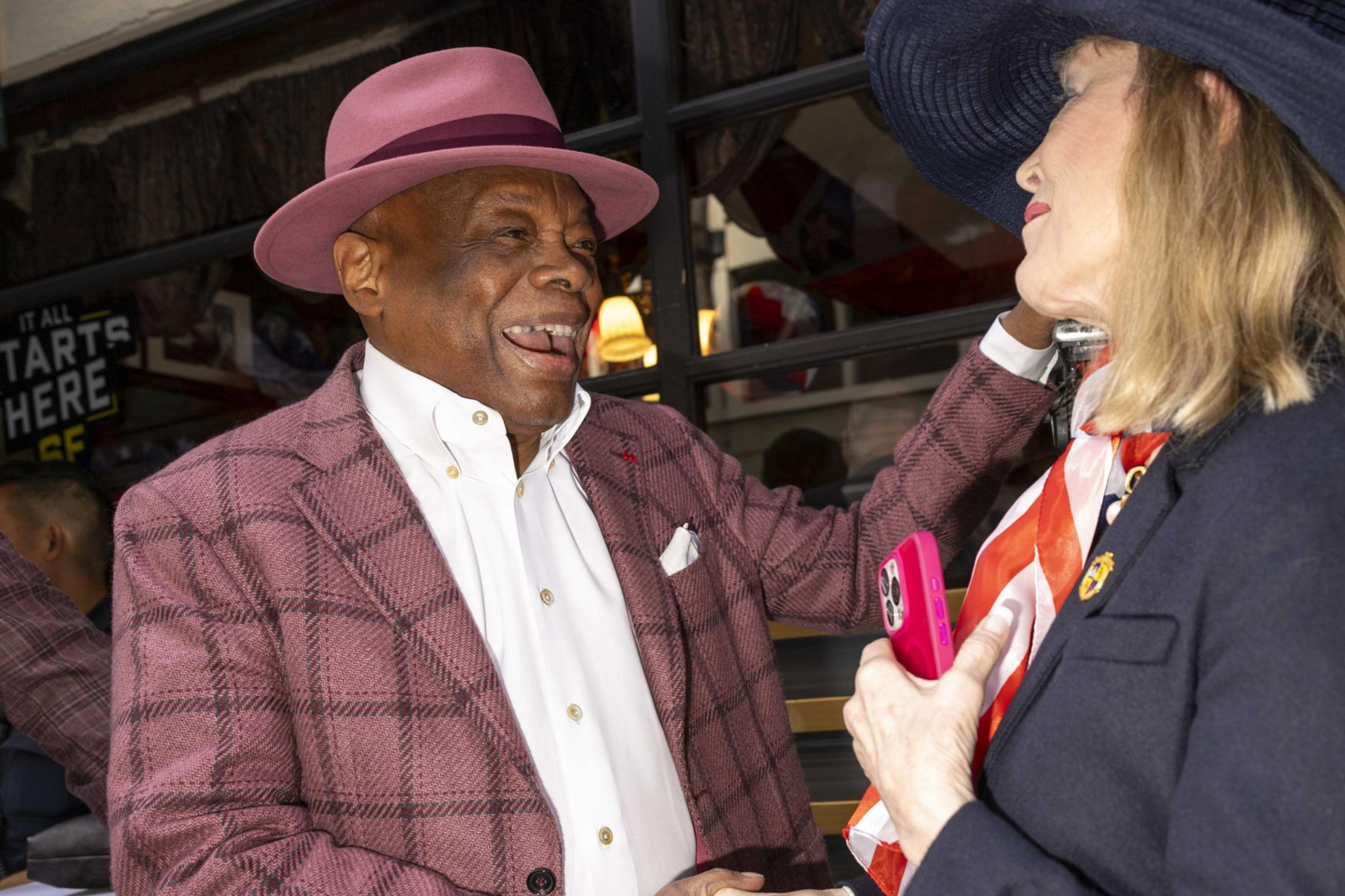 Two people are warmly greeting each other, both wearing stylish hats. The man in a pink hat and checked jacket is smiling broadly, shaking hands with the woman in a blue hat.