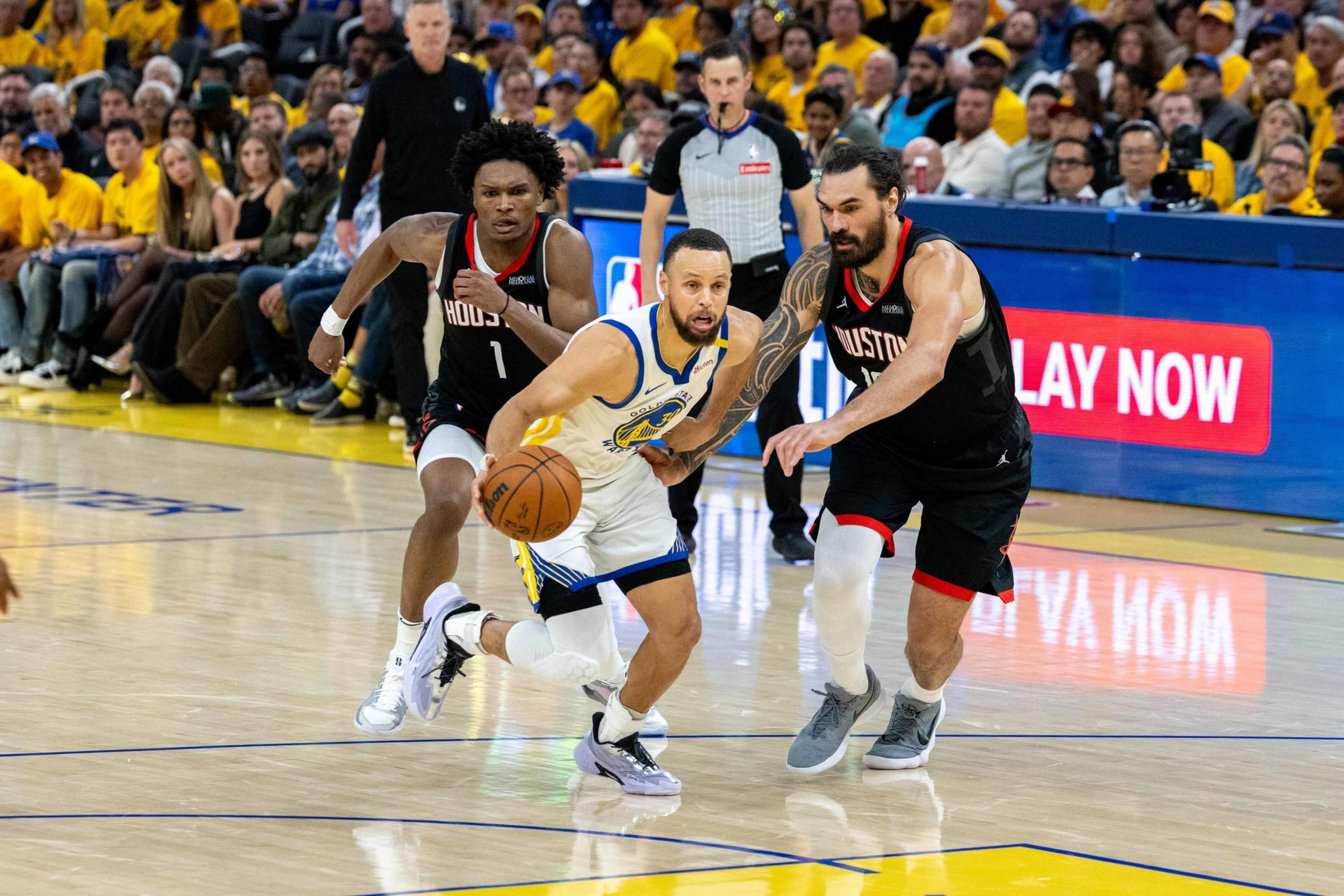 A basketball player in a Warriors uniform dribbles the ball while two opposing players in black Rockets uniforms defend him on a crowded indoor court.