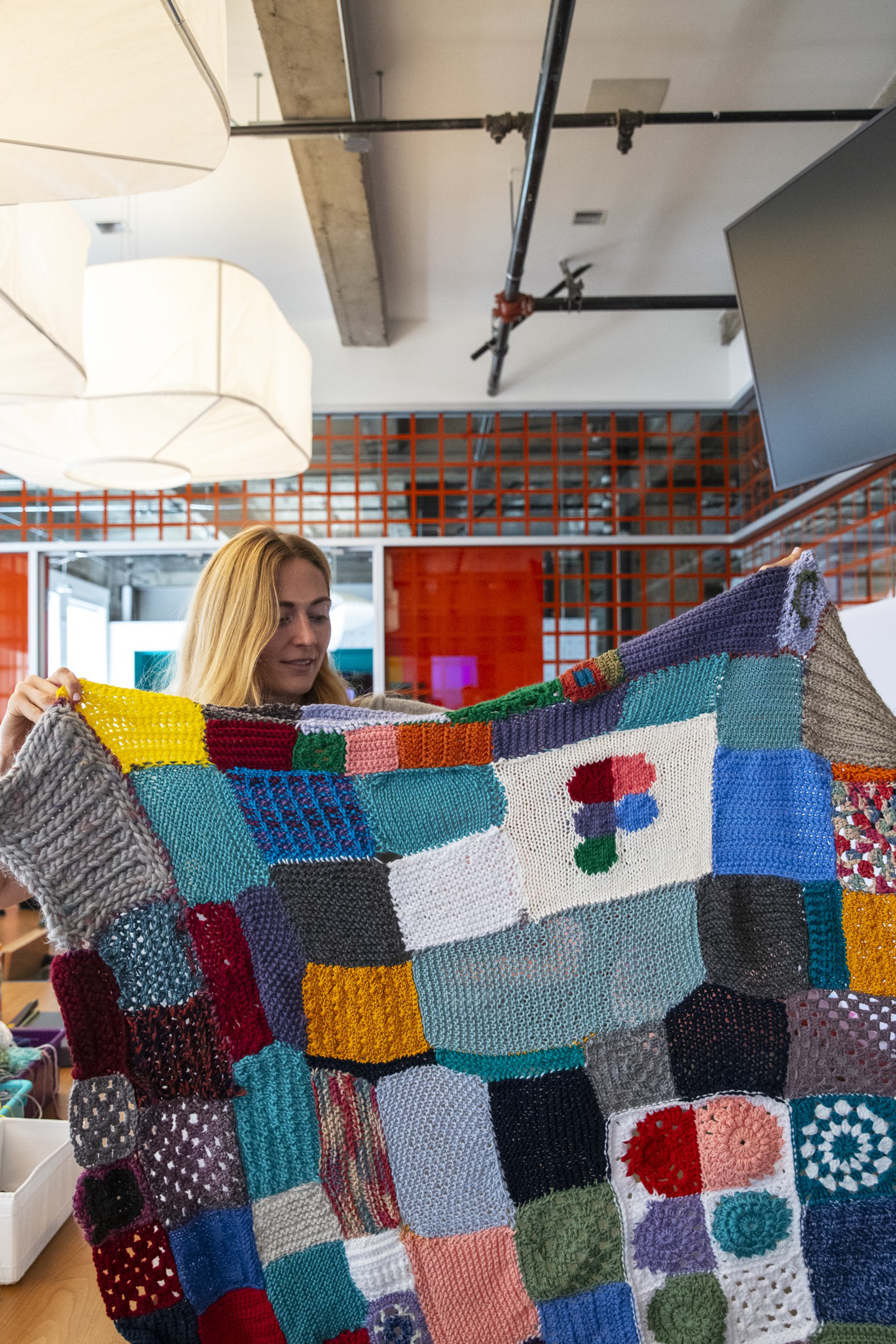 A woman holds up a colorful, patchwork knitted blanket featuring a white square with a multicolored, pixelated "F" logo in the center.