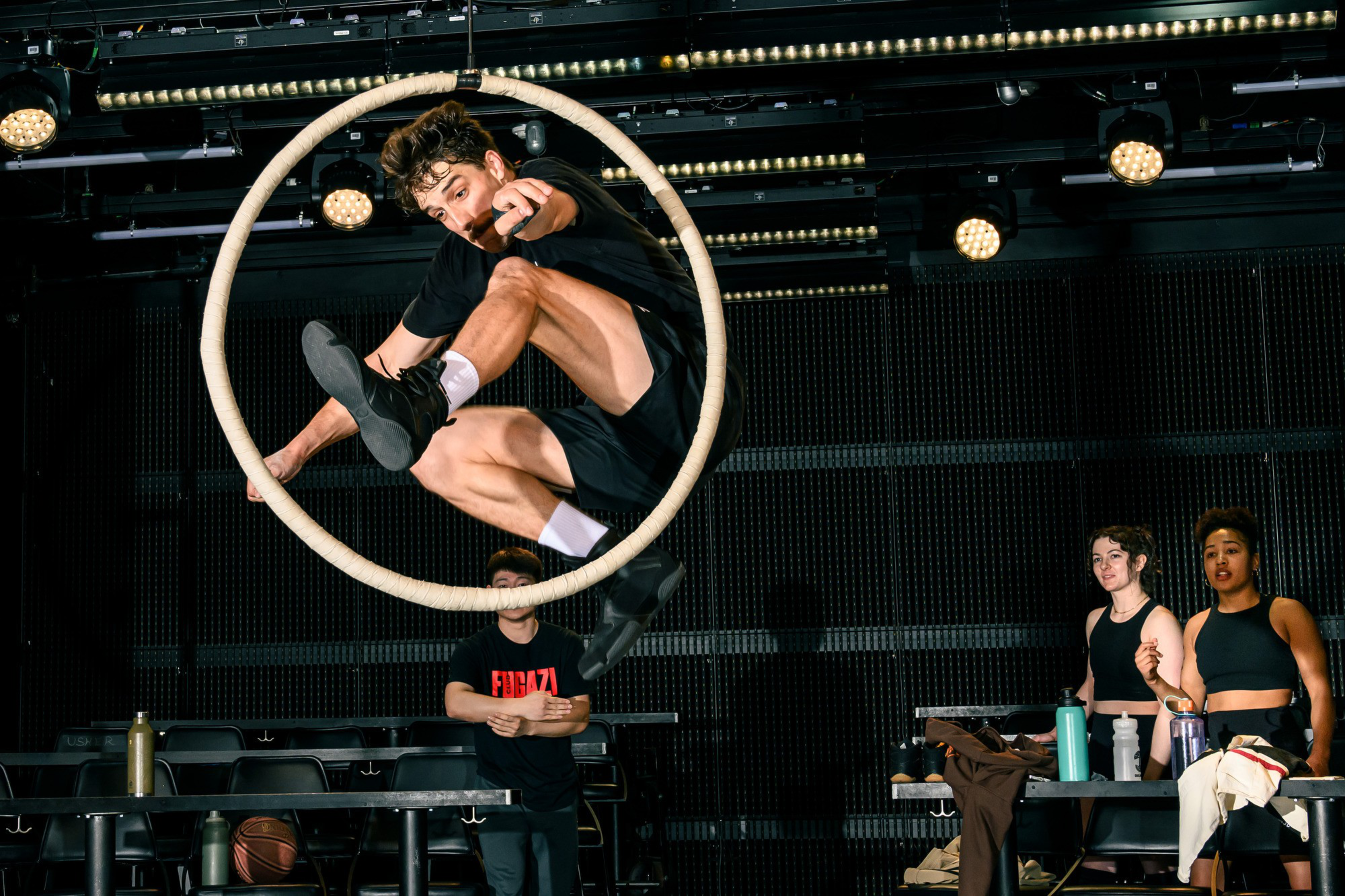 A person is performing a dynamic acrobatic move inside a circular hoop suspended in the air, with three observers in the background on a stage-like setting.