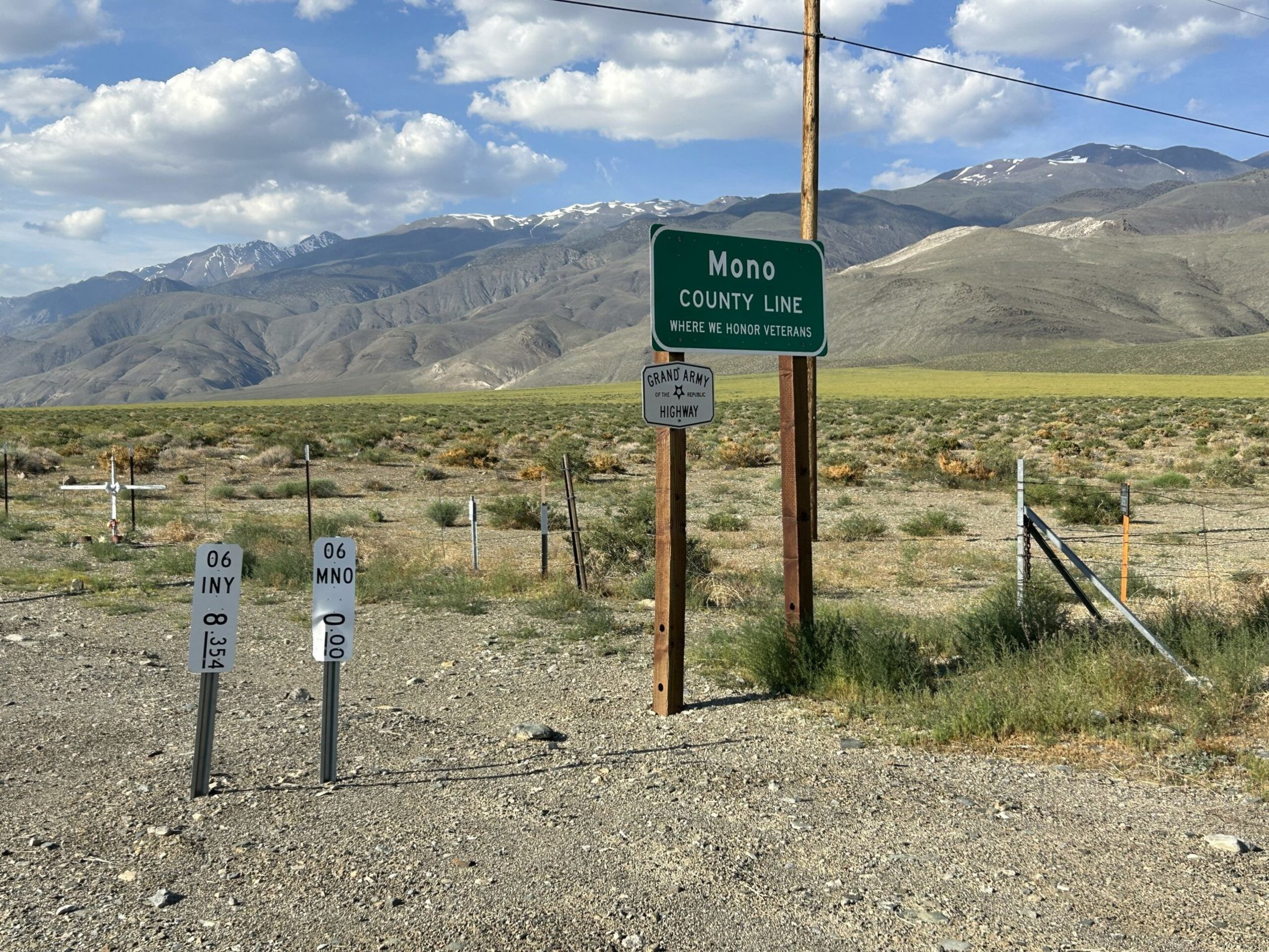 A roadside marks the Mono County line with signs, dry grassland, and distant mountains under a partly cloudy blue sky.