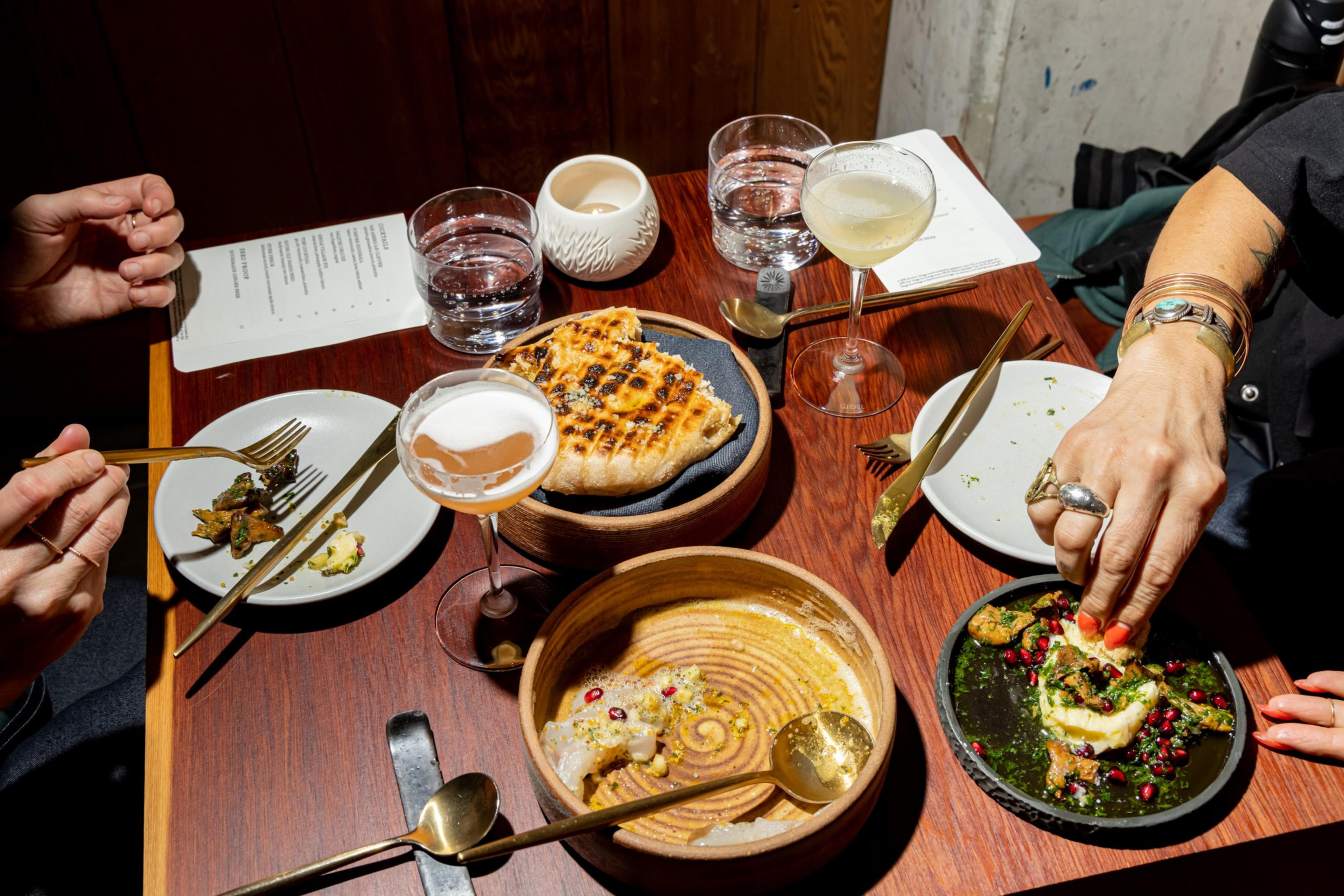 A table with grilled bread, a half-eaten dish with pomegranate seeds, two cocktails, water glasses, and people eating with gold utensils.