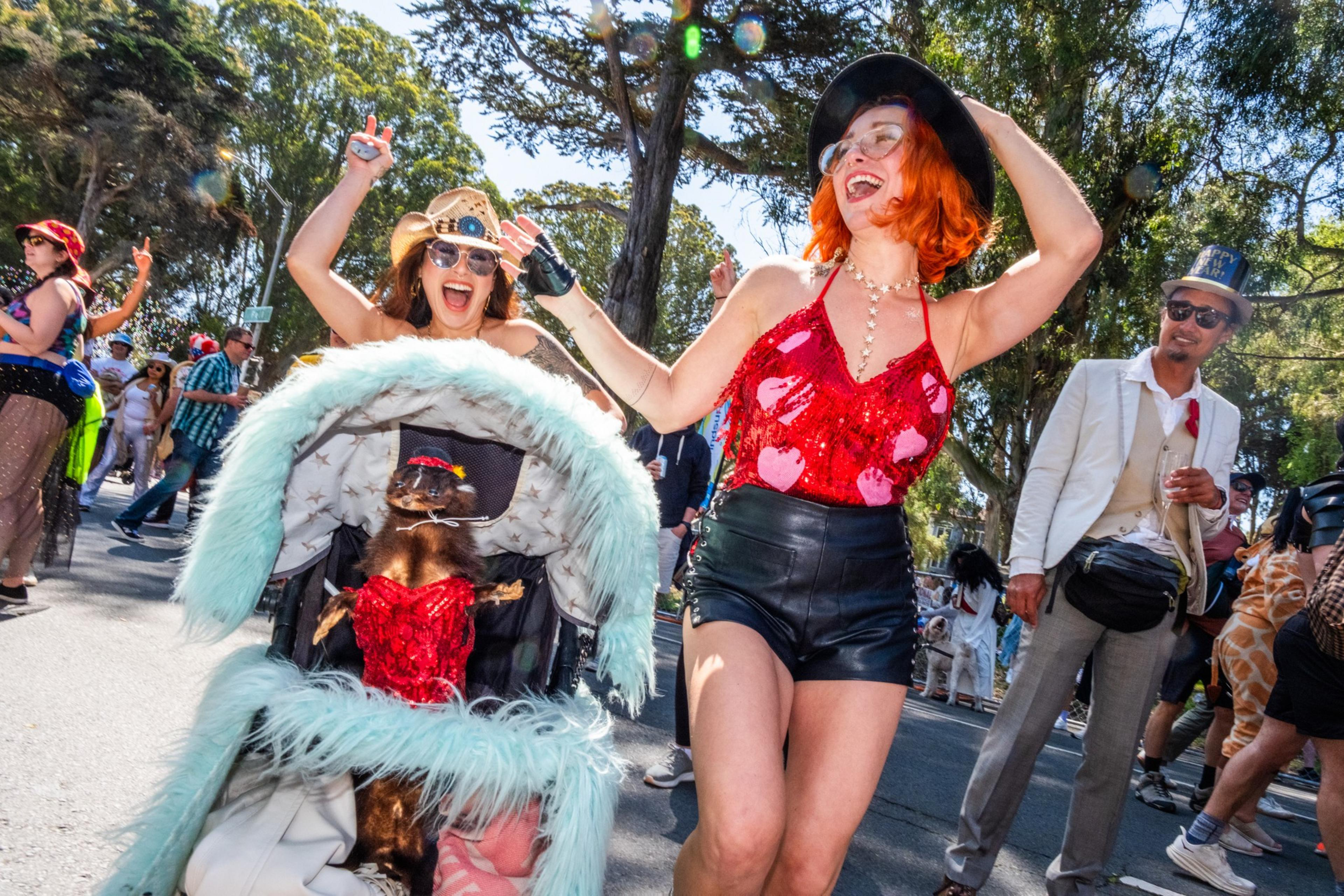 Two women, wearing hats and vibrant outfits, joyfully dance beside a stroller with a dressed-up dog. People enjoy the sunny outdoor scene around them.