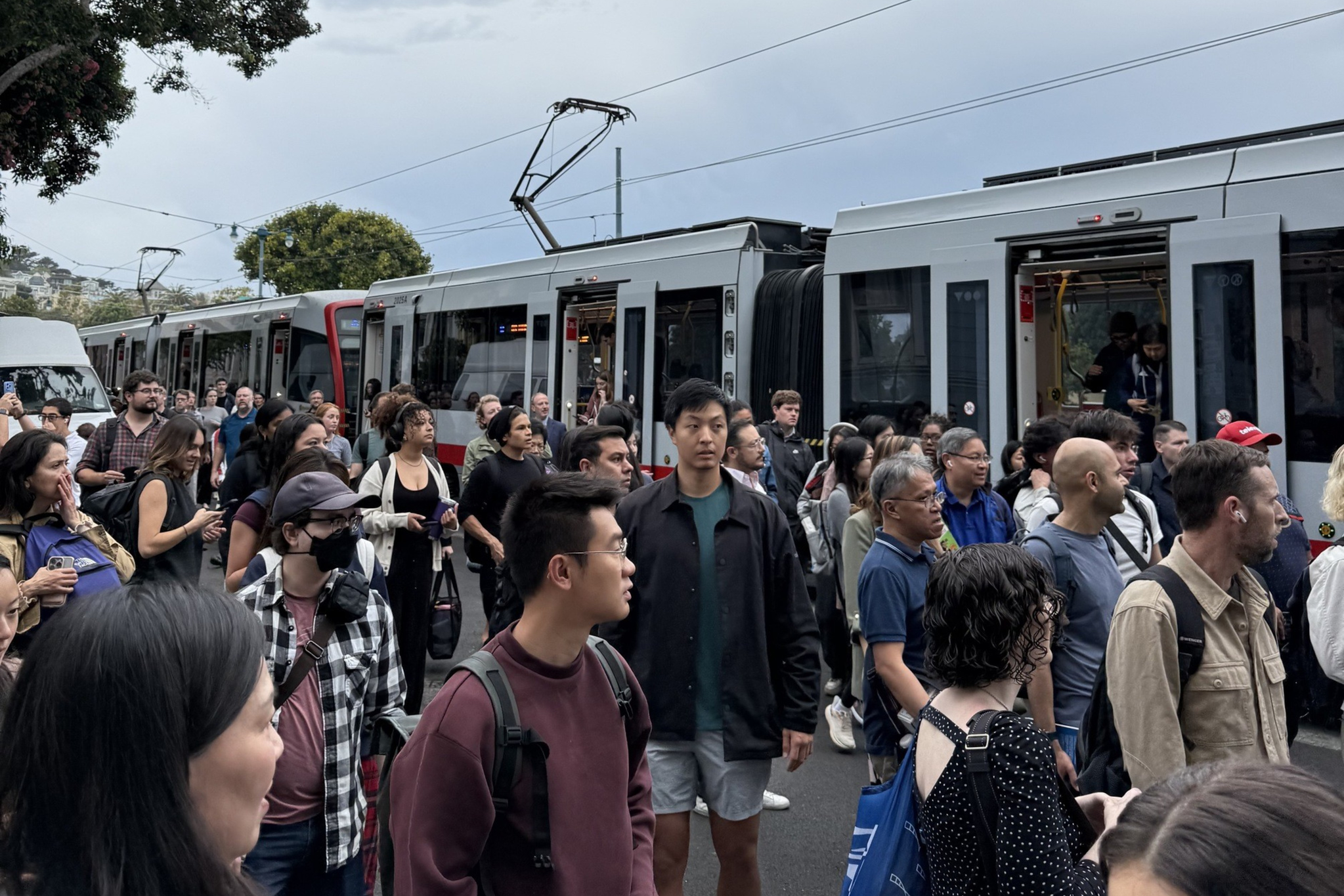 A large crowd of people stands on a street near a stationary tram, many looking ahead or talking, under an overcast sky with trees in the background.