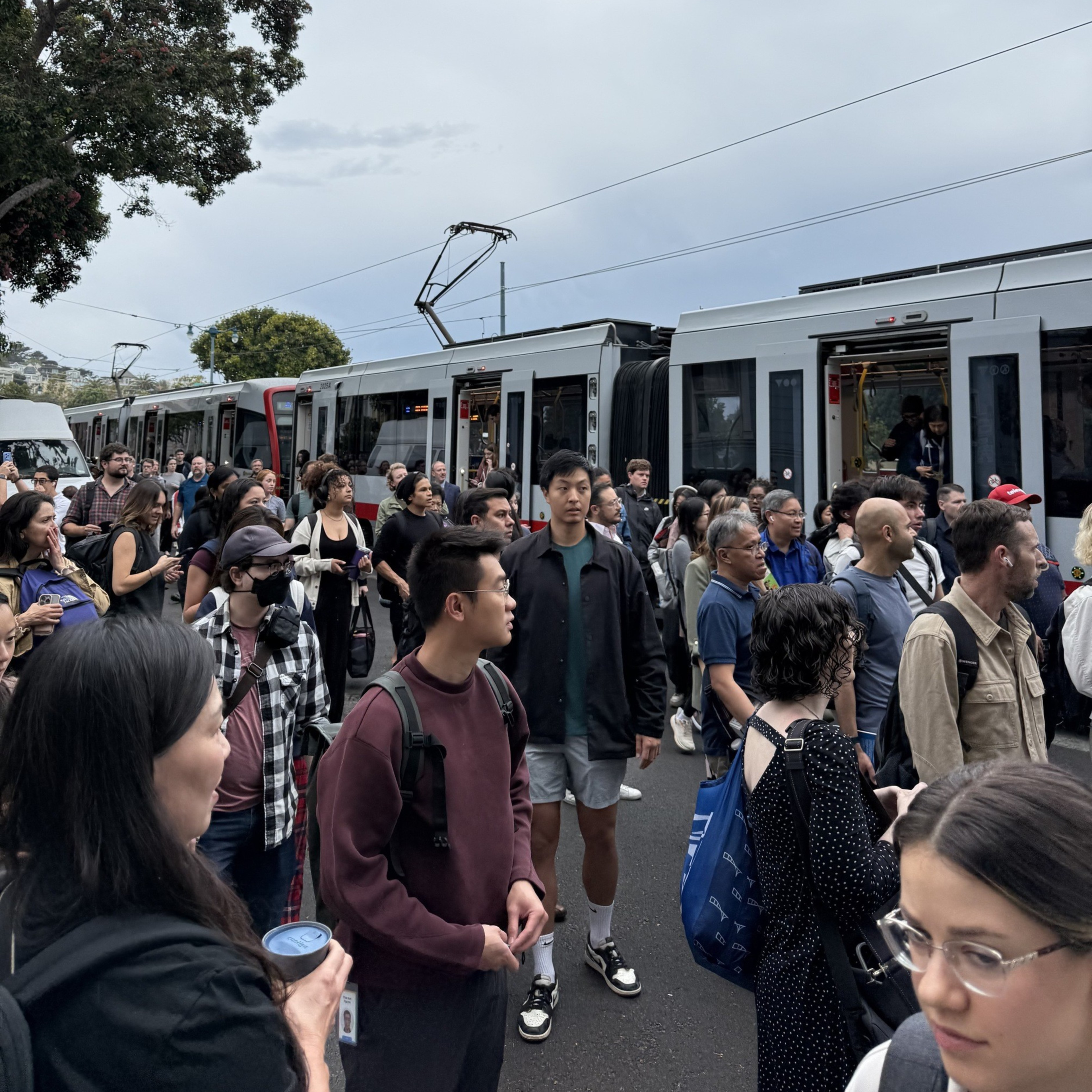 A large crowd of people stands on a street near a stationary tram, many looking ahead or talking, under an overcast sky with trees in the background.