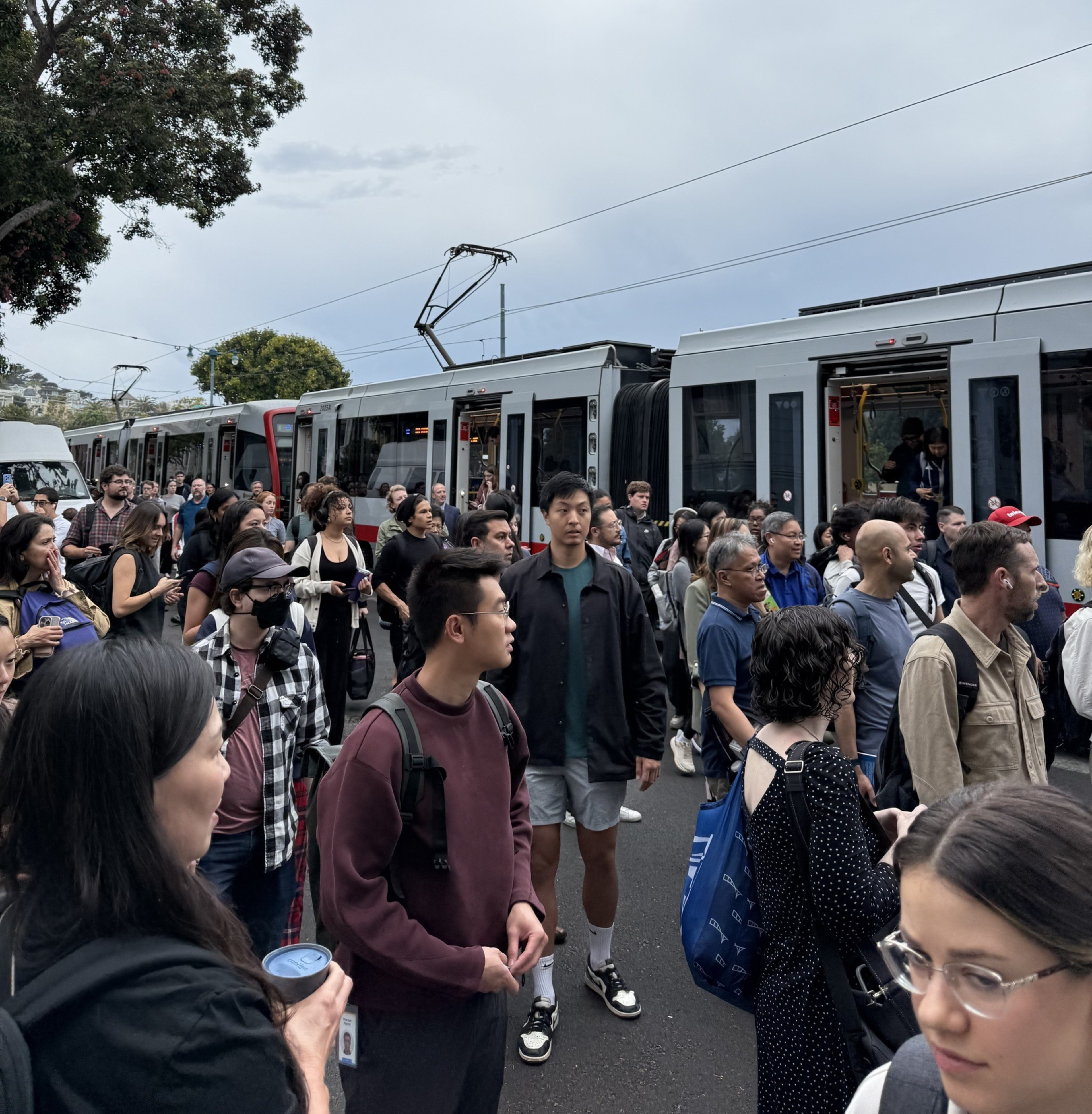 A large crowd of people stands on a street near a stationary tram, many looking ahead or talking, under an overcast sky with trees in the background.