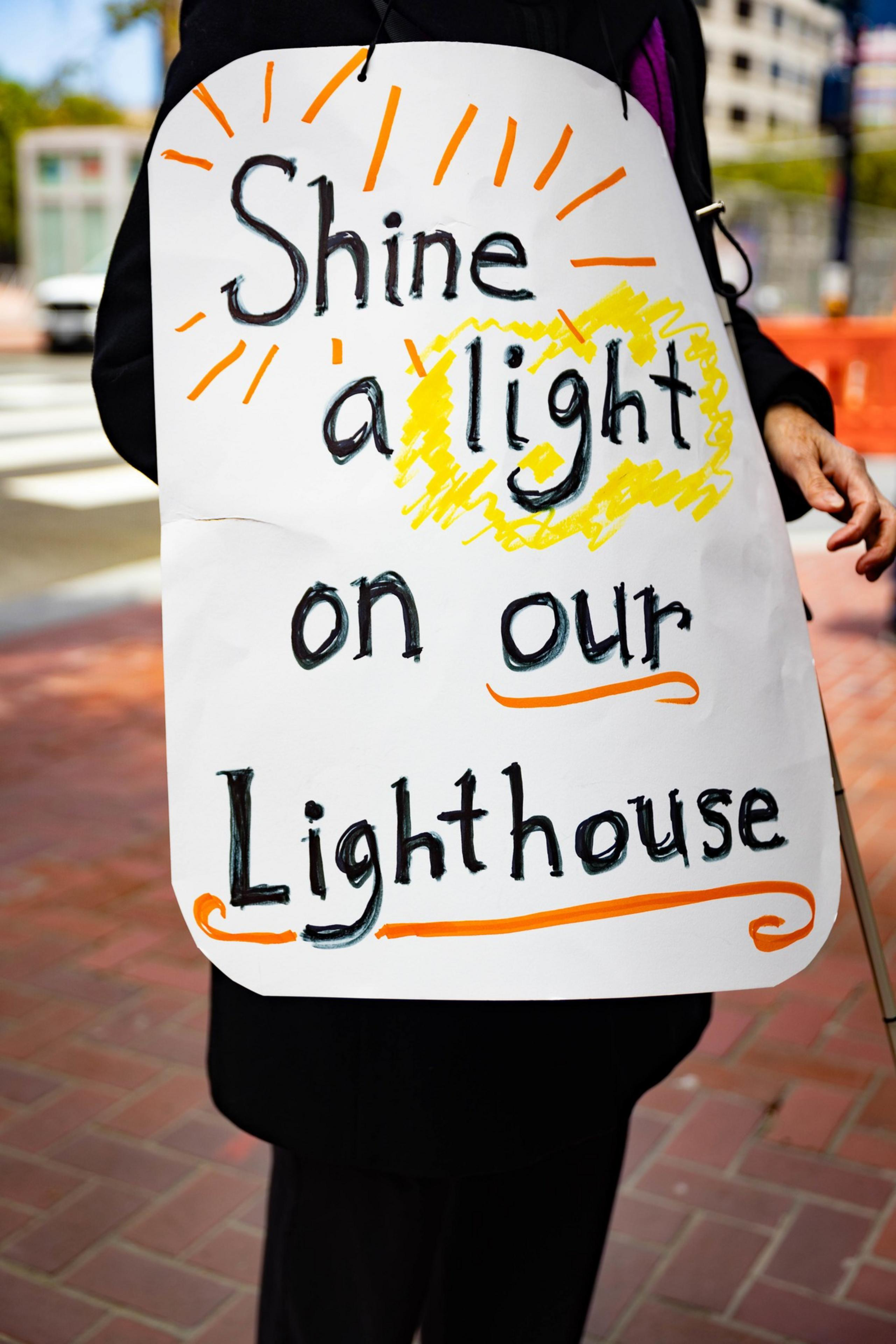 The image shows a hand-held sign with the words &quot;Shine a light on our Lighthouse,&quot; decorated with orange and yellow highlights.