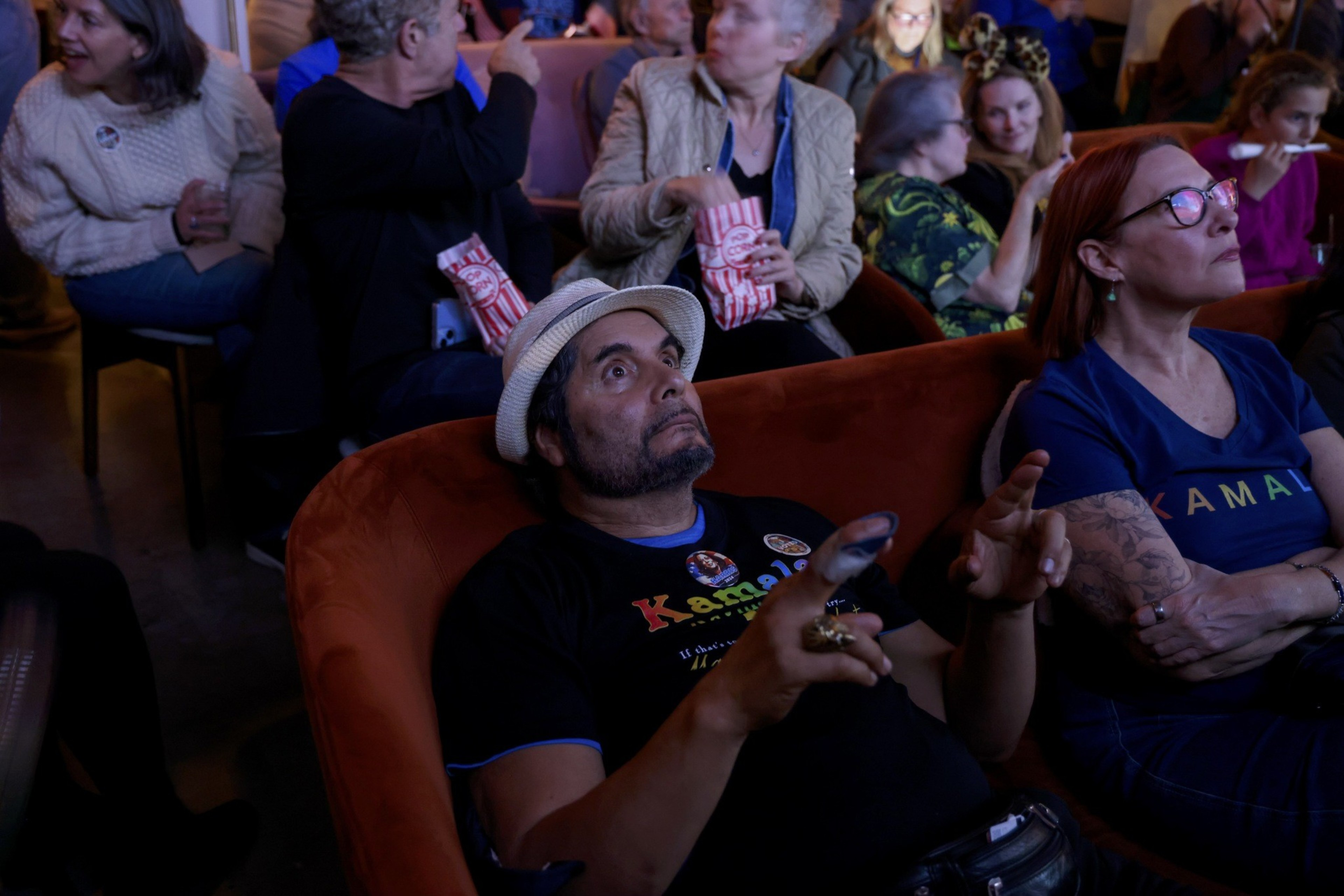 A man in a hat sits on an orange chair, holding a phone with his finger bandaged, while people around him chat and eat popcorn in a crowded, dimly lit room.