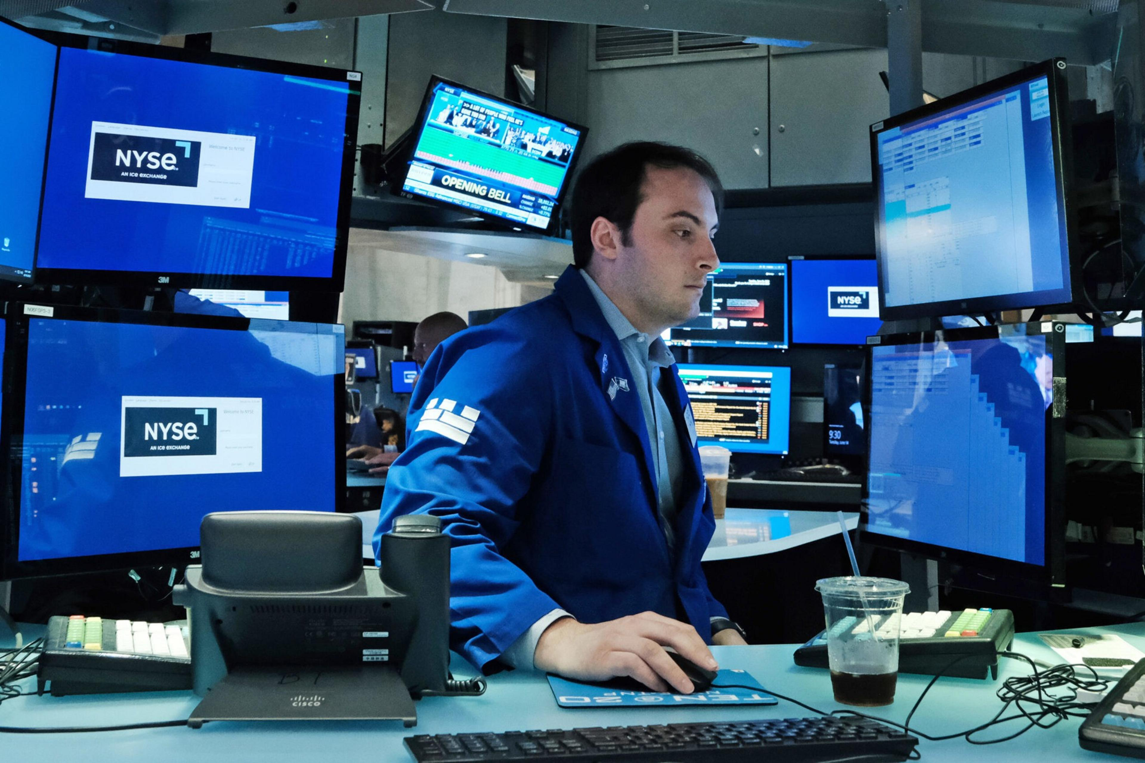 A man in a blue jacket works at a desk surrounded by multiple computer monitors displaying NYSE stock market data.