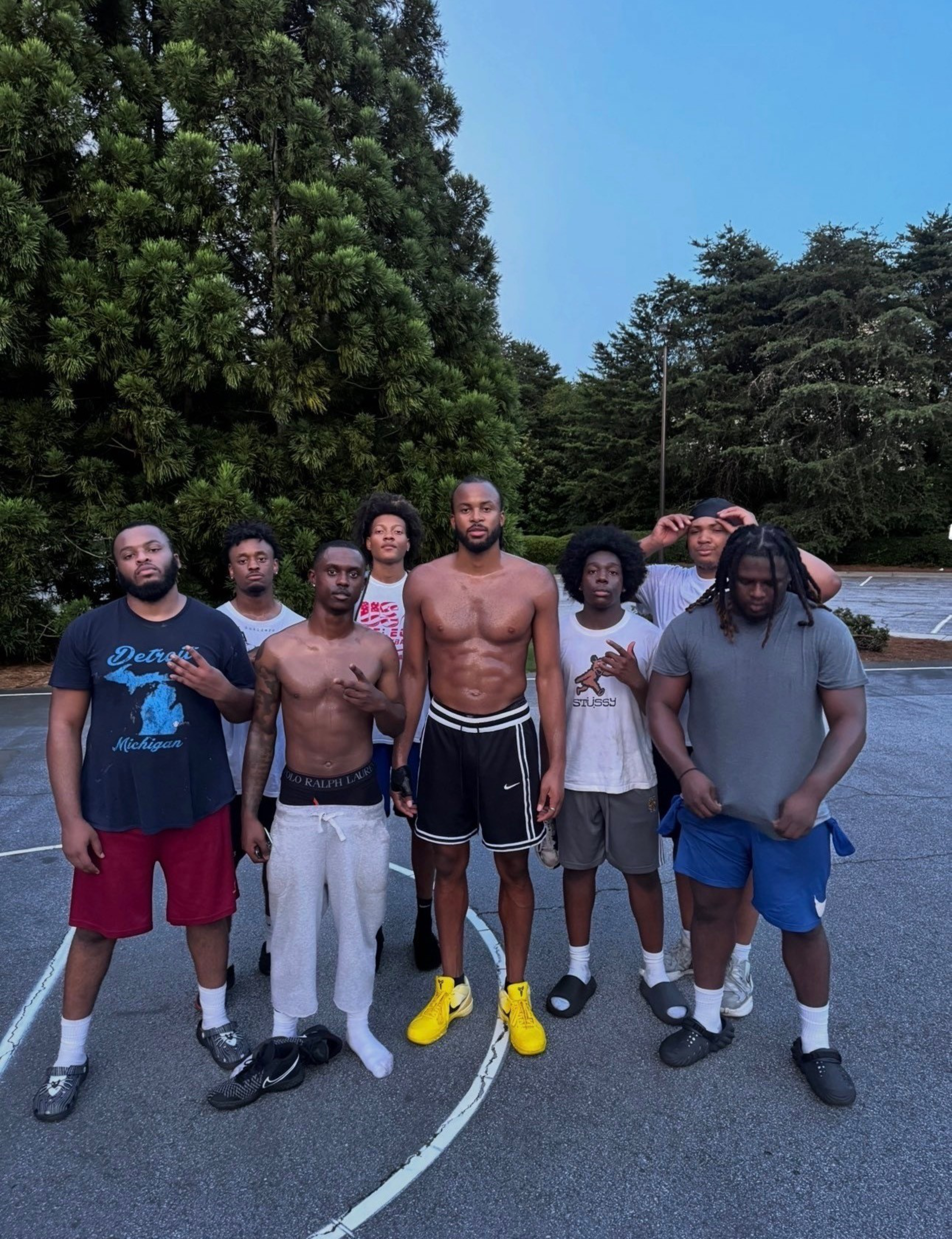 Eight young men stand on an outdoor court, some shirtless, wearing casual athletic clothing and sneakers, with tall green trees and a clear sky behind them.