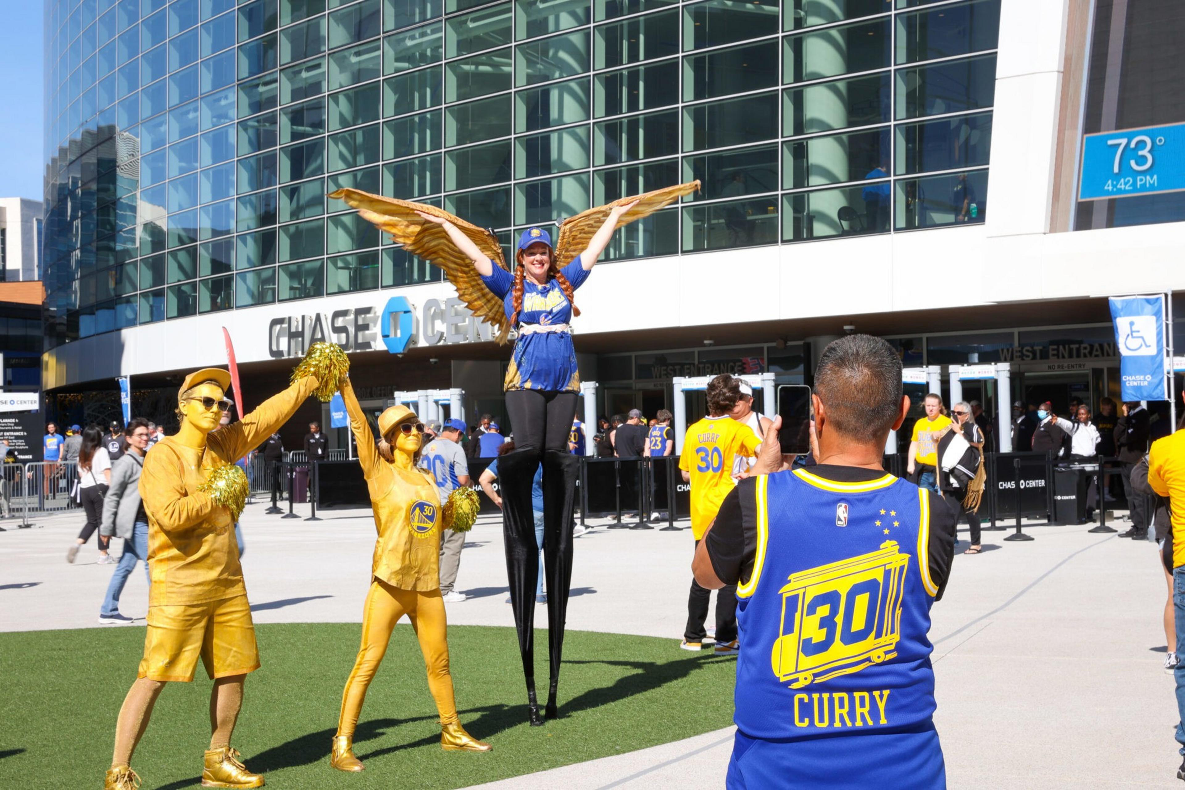 A man wearing a Curry basketball jersey takes a photo of two gold-painted cheerleaders and a performer on stilts with golden wings outside Chase Center.