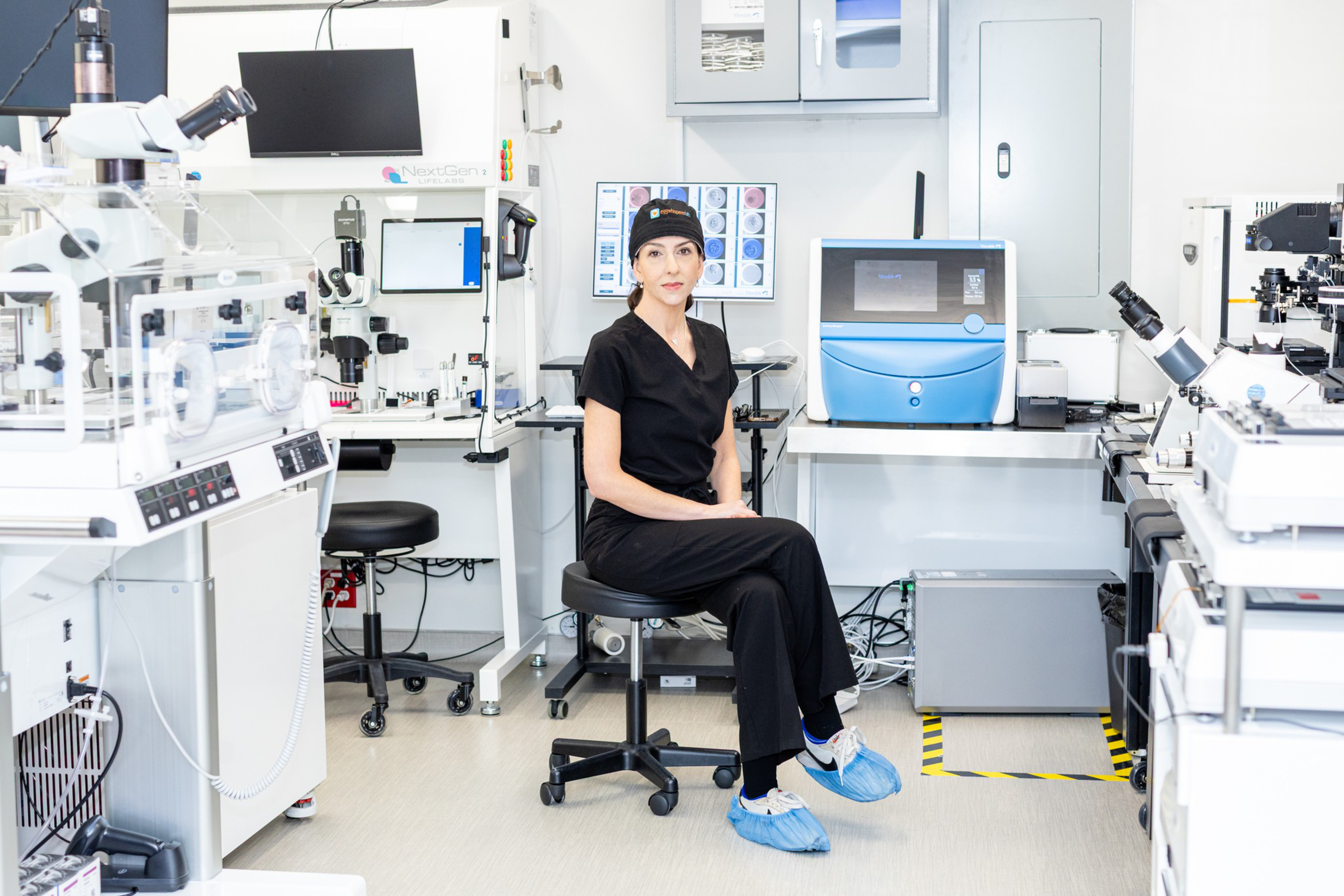 A woman in black scrubs and shoe covers sits on a stool in a bright laboratory filled with microscopes, monitors, and scientific equipment.
