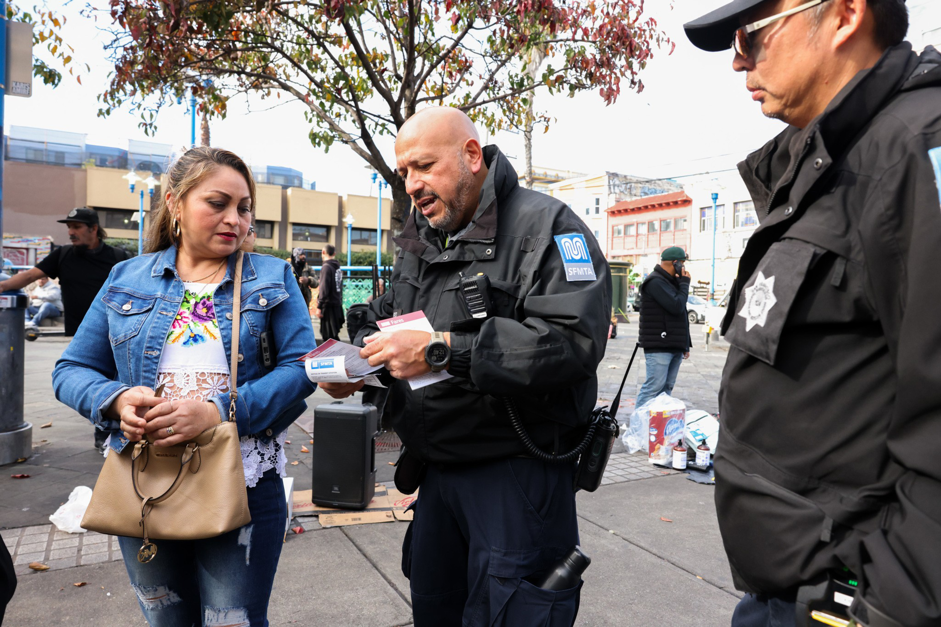 A woman with a handbag stands in an outdoor area as two uniformed officers, surrounded by people, speak with her and show her papers.