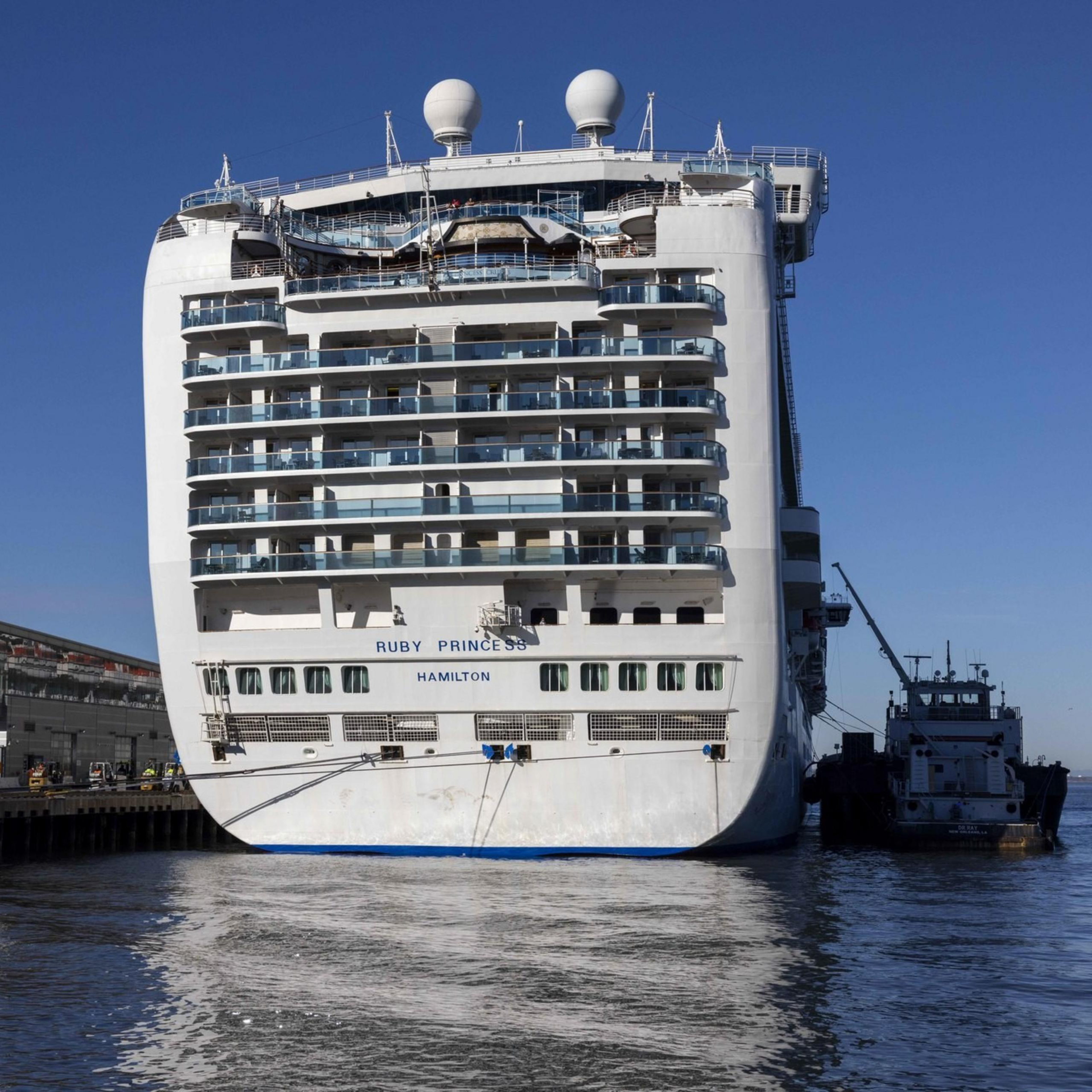A large cruise ship is docked at a port, with buildings visible to the left and a small boat alongside it. The sky is clear and blue.