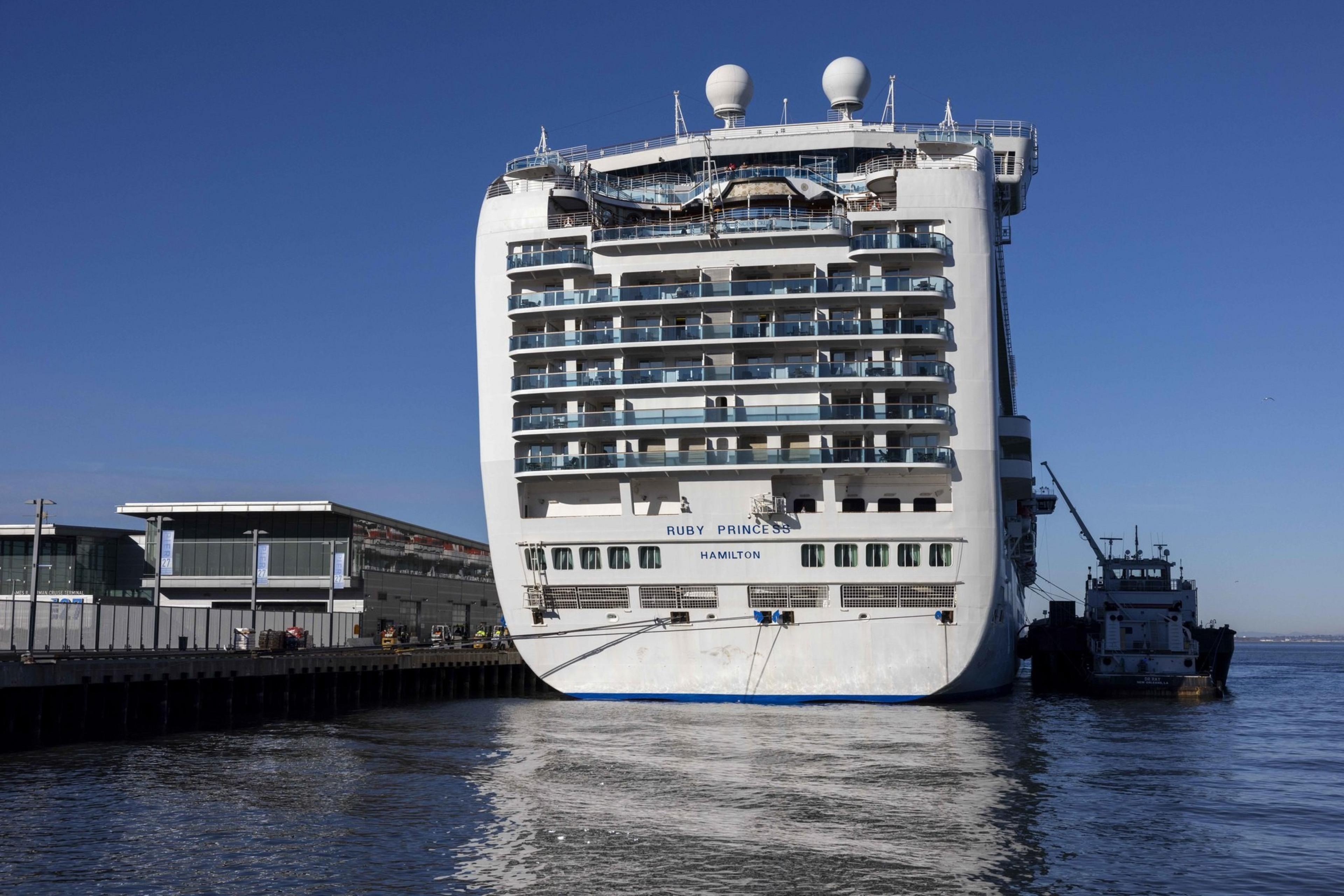 A large cruise ship is docked at a port, with buildings visible to the left and a small boat alongside it. The sky is clear and blue.