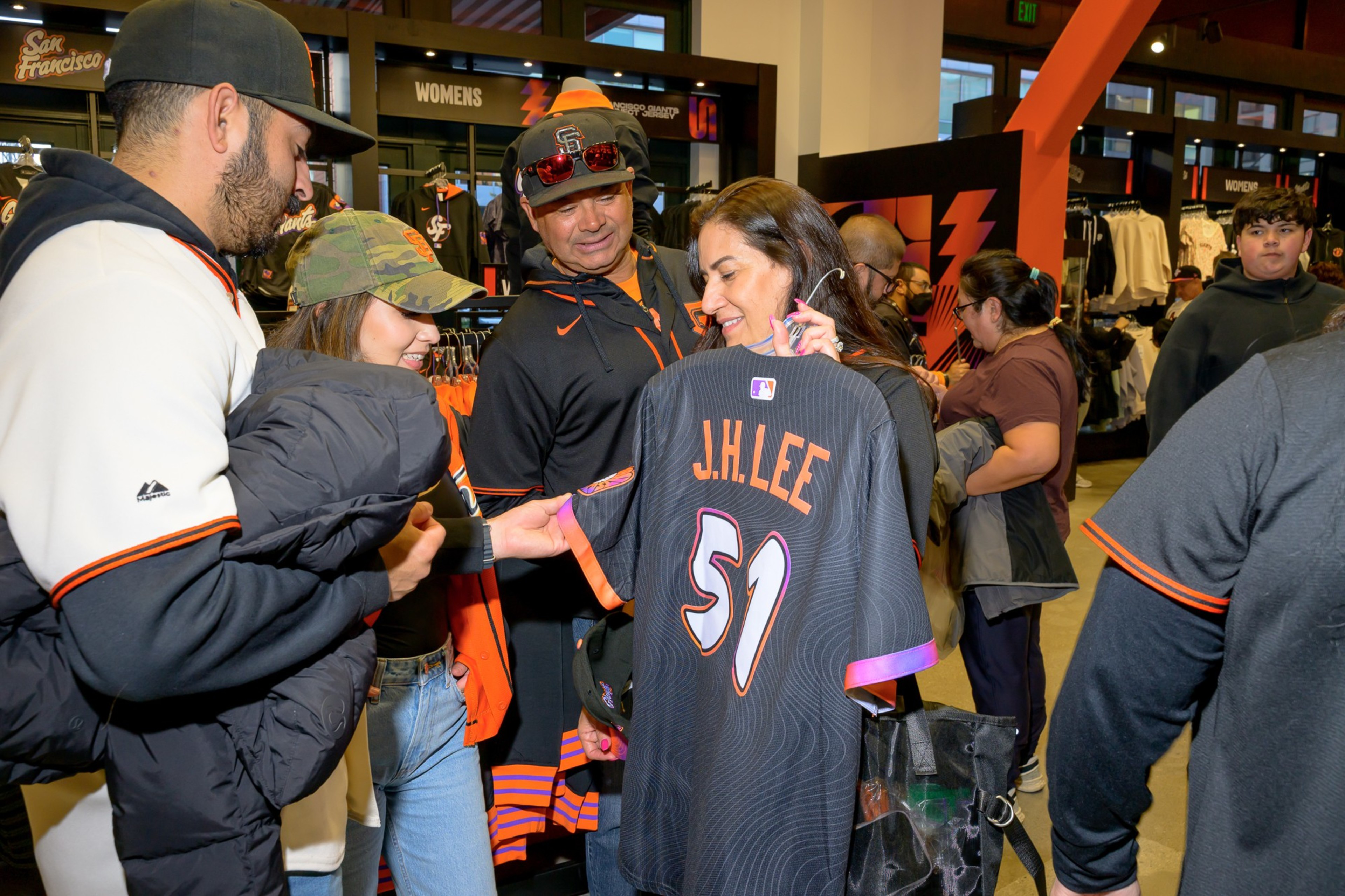 A group of people are shopping at a sports merchandise store. A woman holds up a black baseball jersey with &quot;J.H. Lee&quot; and the number 51 on the back.