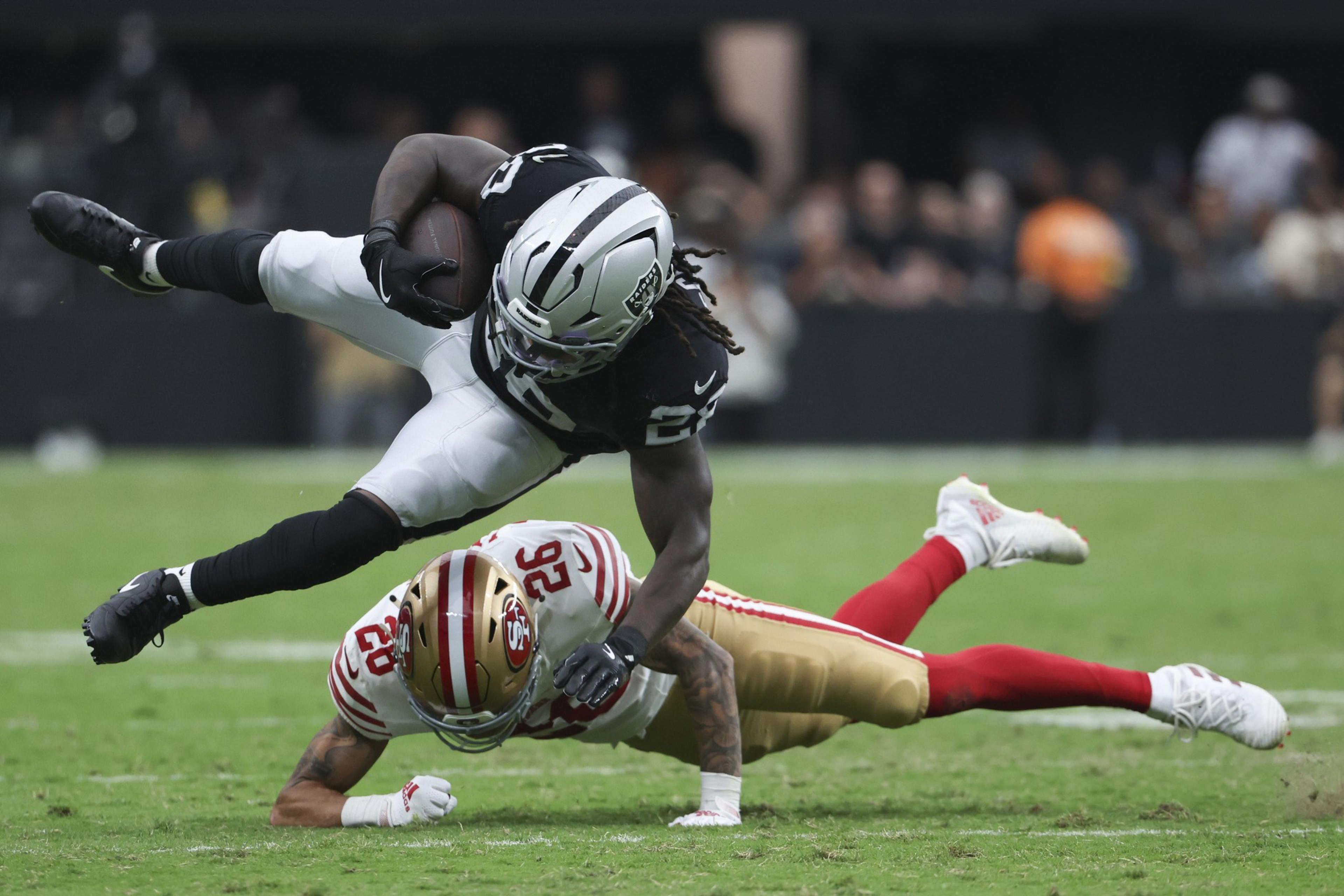A football player in a black and white uniform leaps over a player in a red and gold uniform who is diving forward on the field.