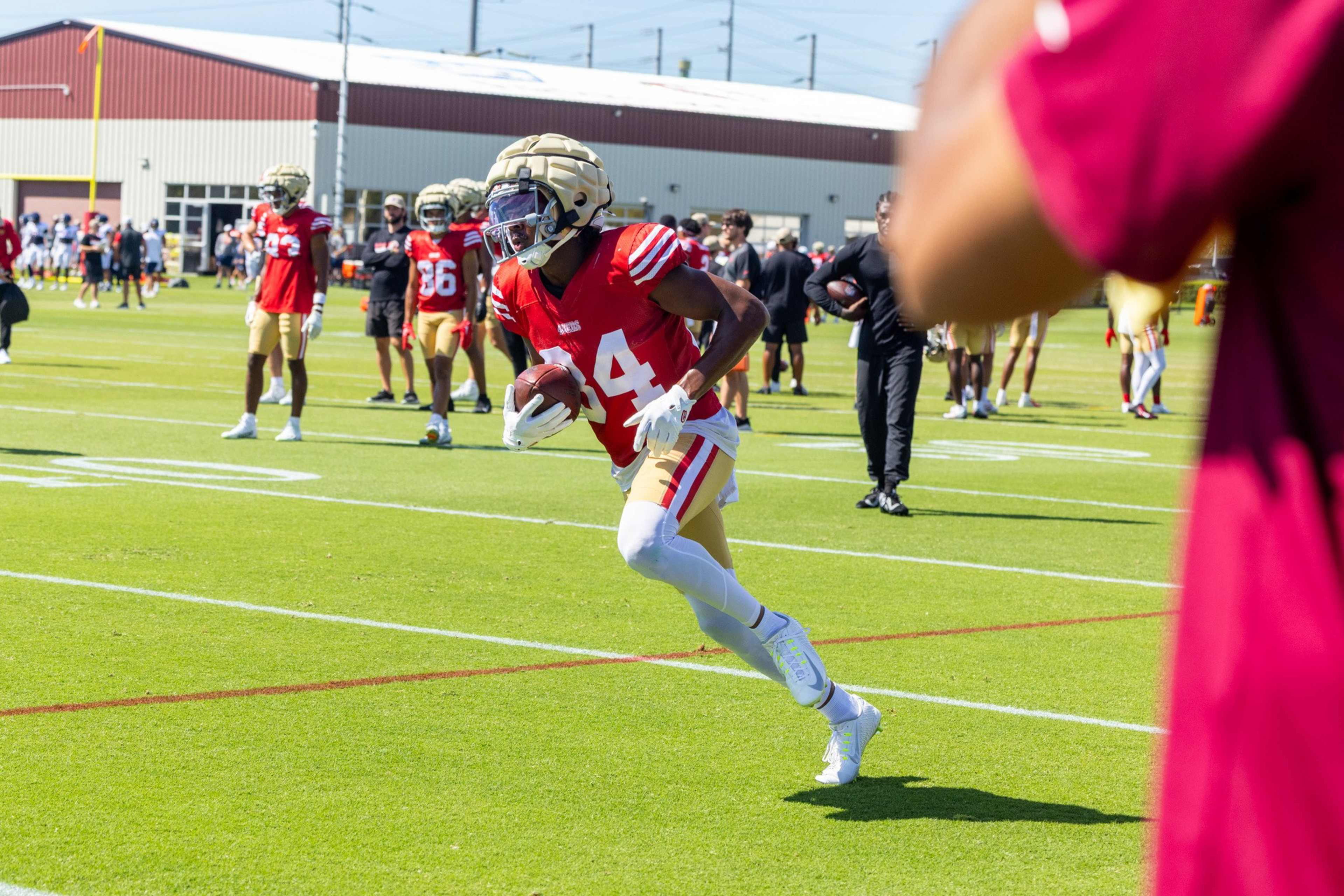 A football player in a red jersey with number 14 runs on a field holding a football, while teammates and coaches watch from behind.
