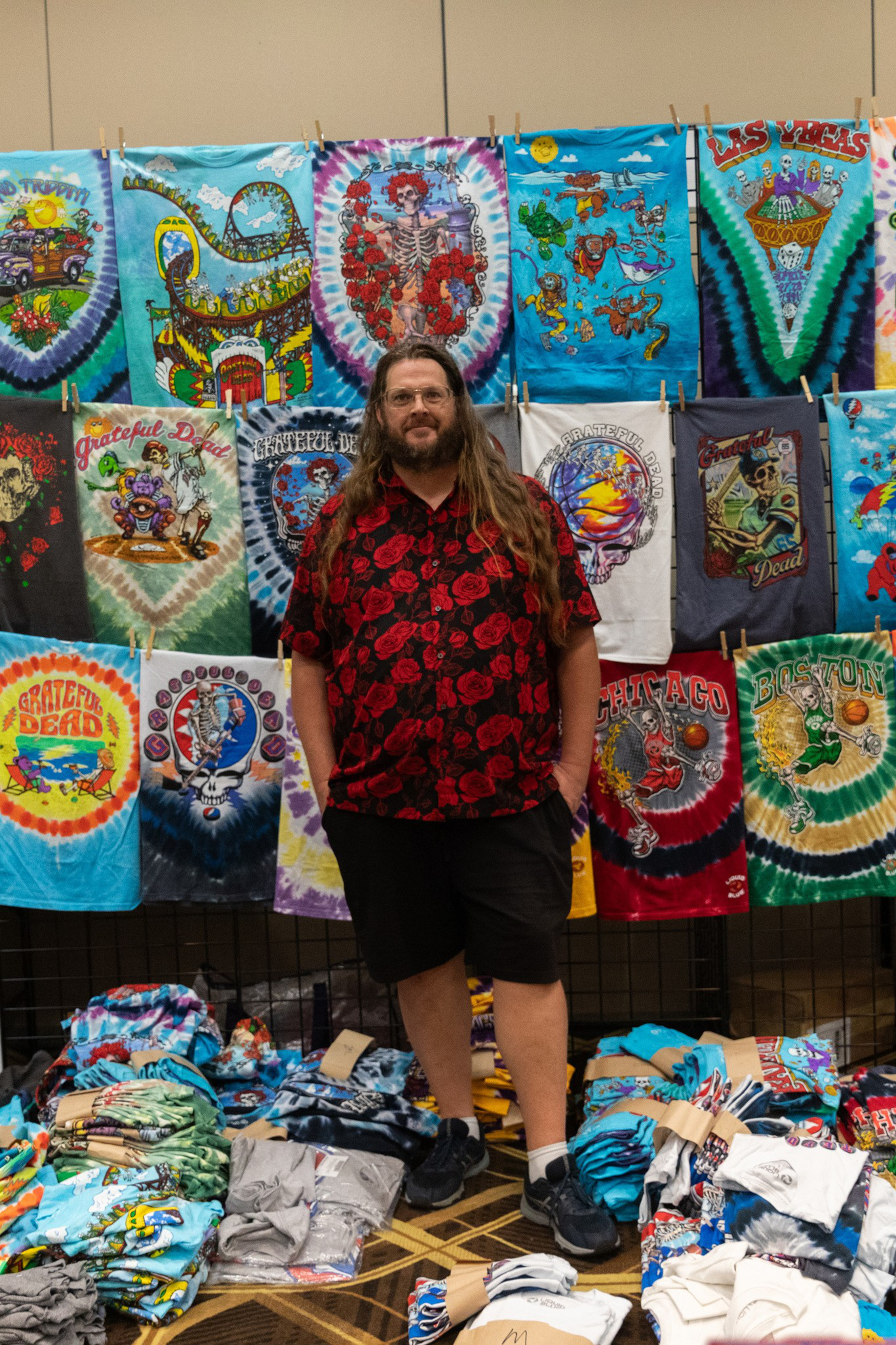 A person stands in front of a colorful display of tie-dye and graphic T-shirts, with more shirts piled on the floor. They wear a red rose-patterned shirt and black shorts.