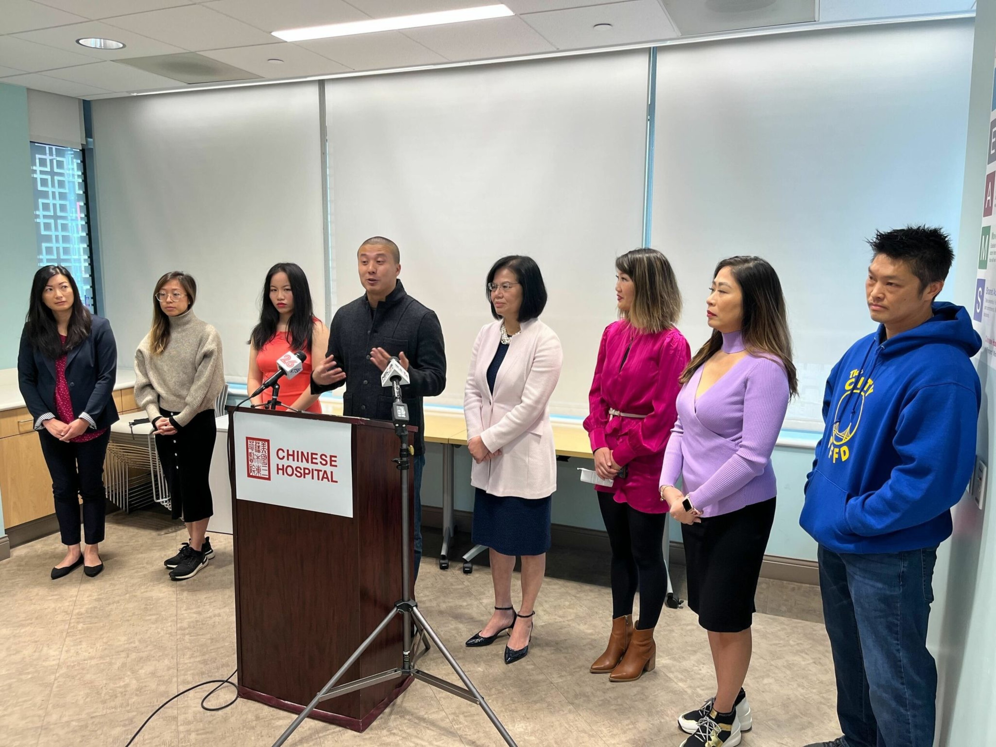 A group of eight people stands in a line behind a podium labeled "Chinese Hospital," with one man speaking into microphones.