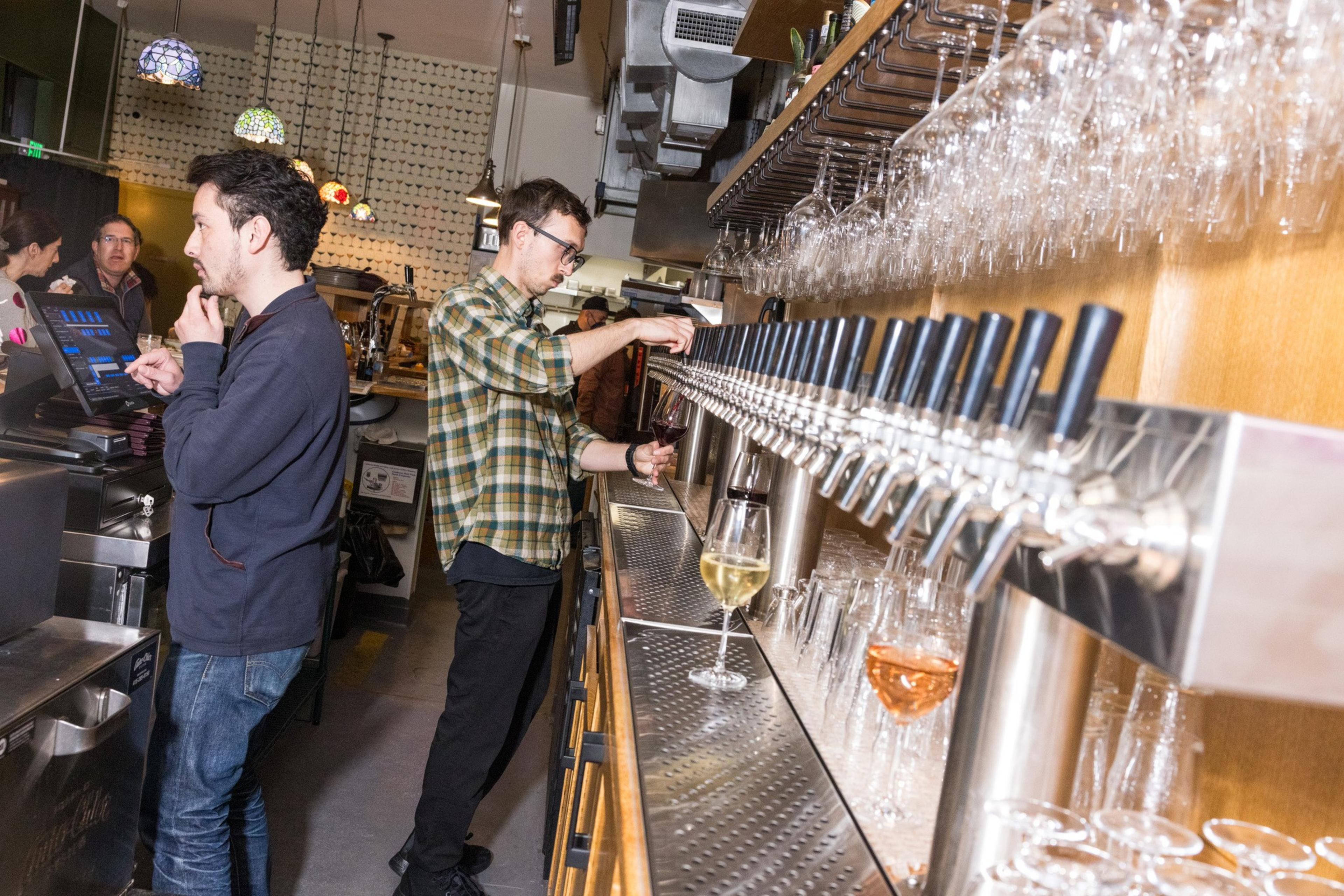 Two people are behind a bar. One is using a touch screen, and the other is pouring drinks from multiple taps. Glasses and bottles are visible.