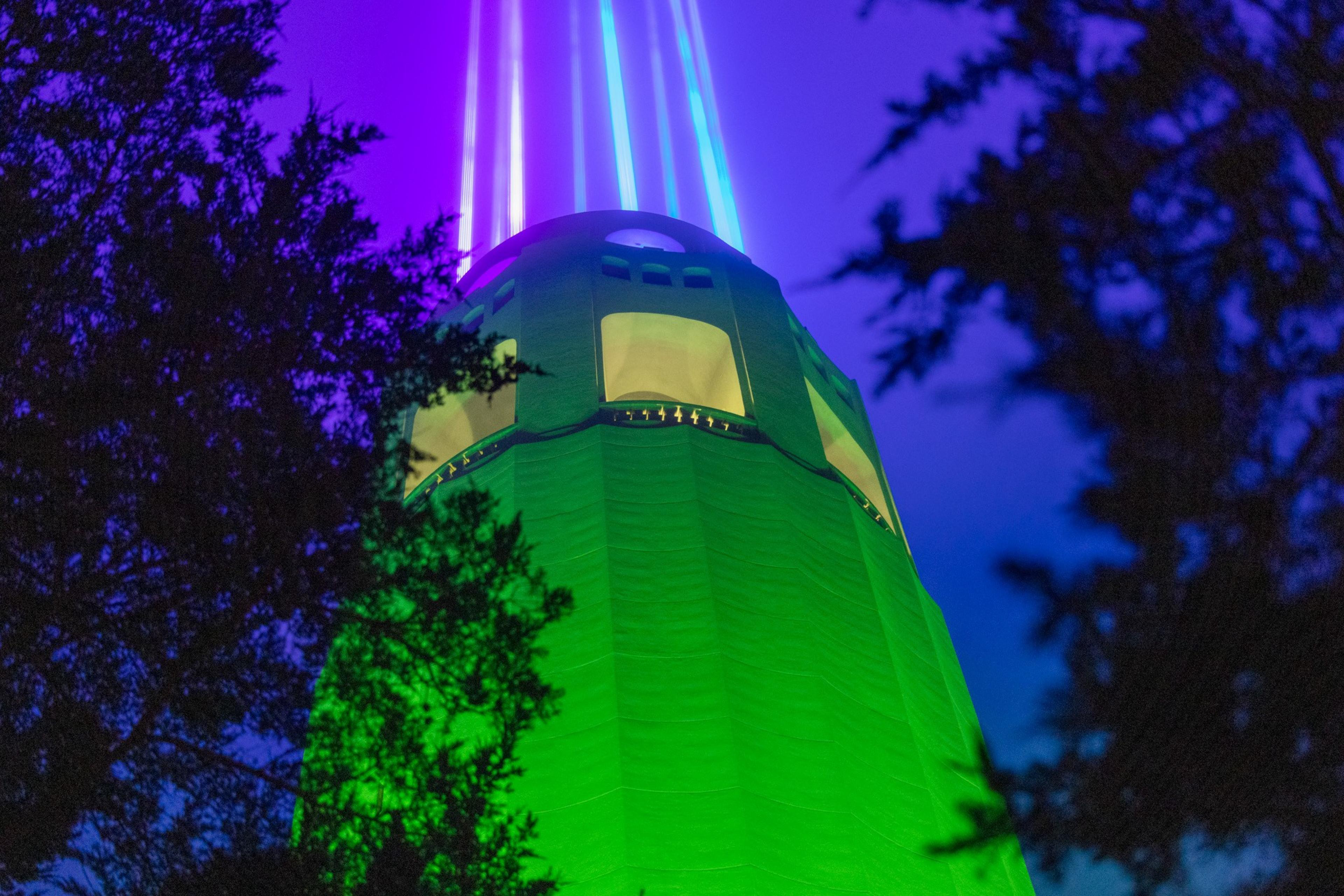 A tower illuminated in green and surrounded by trees shoots vibrant purple and blue light beams into the night sky.