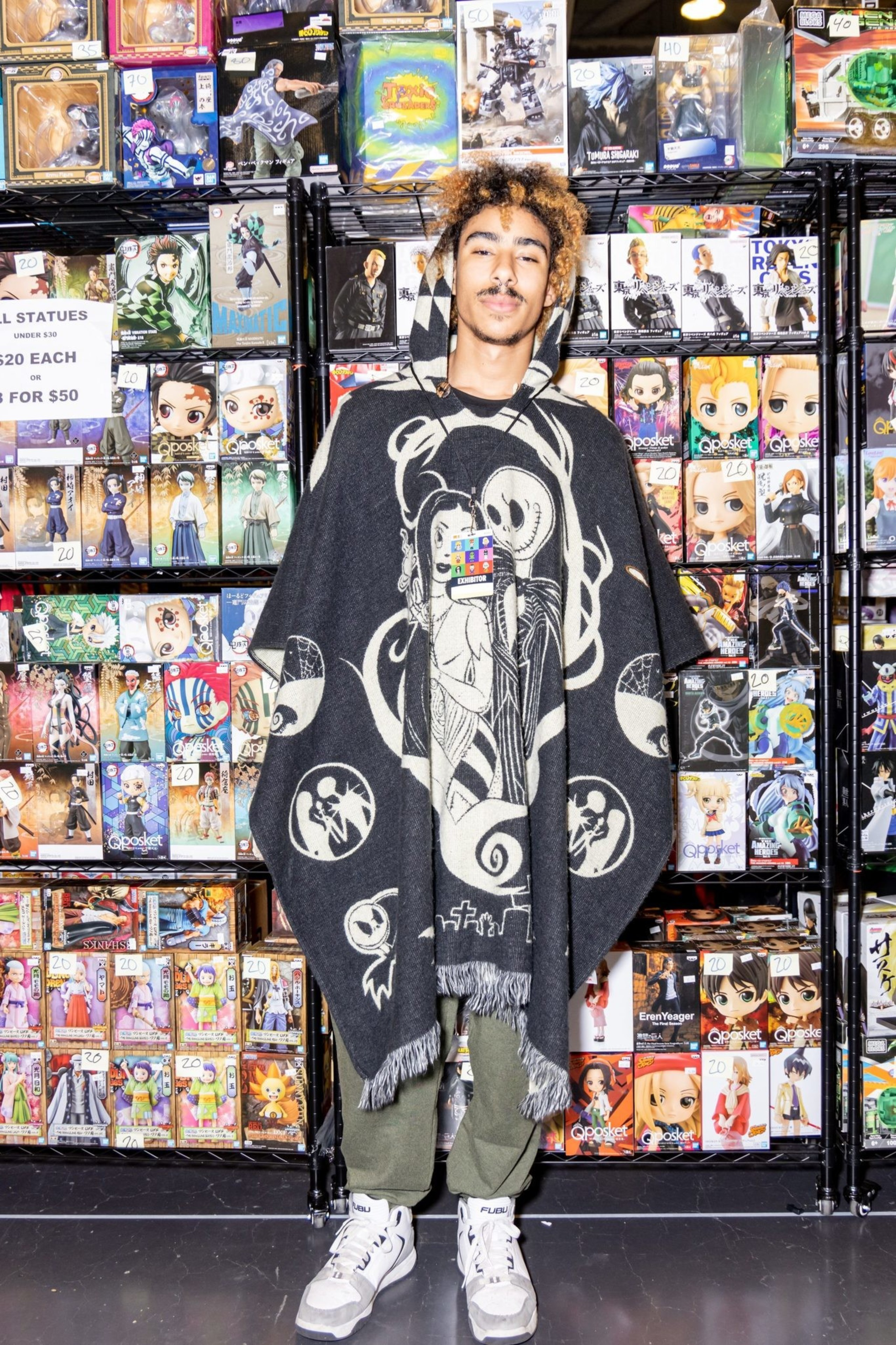 A person stands in front of shelves filled with boxed anime and game figures, wearing a black and white poncho with a spooky design. A sign reads &quot;All Statues $20 Each or 3 for $50.&quot;