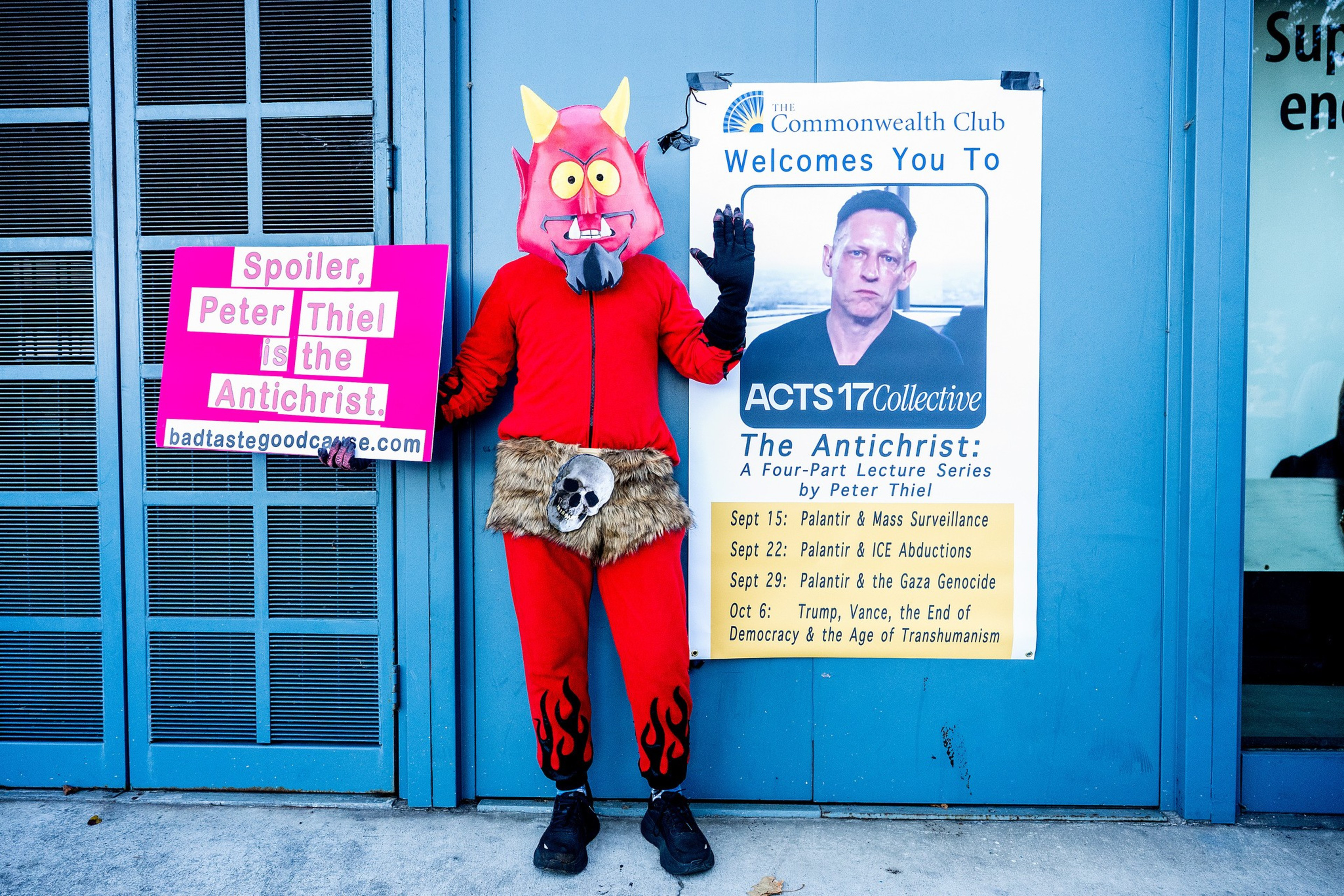 A person in a red devil costume holds a pink sign stating “Spoiler, Peter Thiel is the Antichrist” next to a poster for a lecture series titled “The Antichrist.”