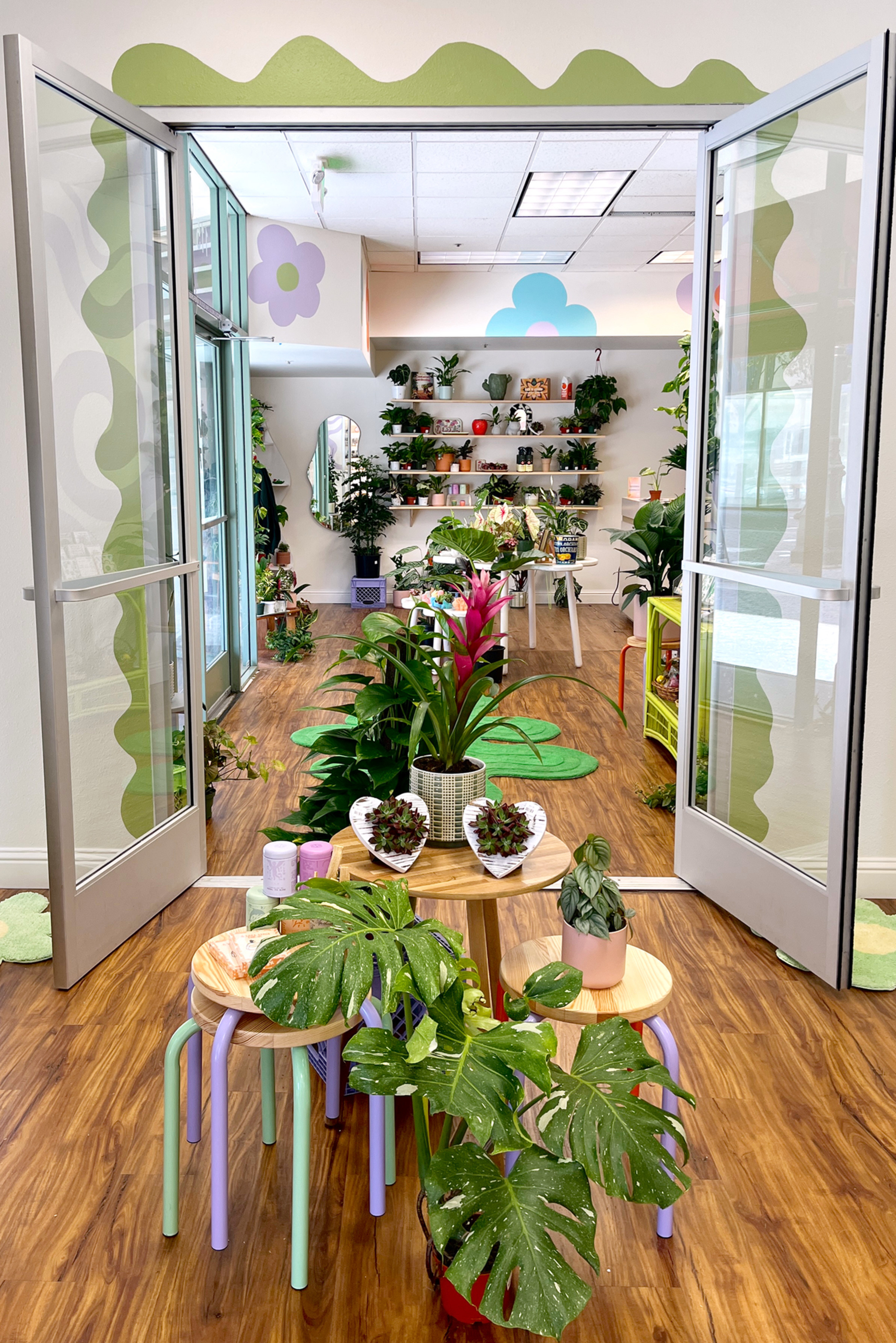 A plant shop with open doors shows vibrant green plants on colorful tables and shelves, set against a backdrop of wooden floors and pastel wall designs.