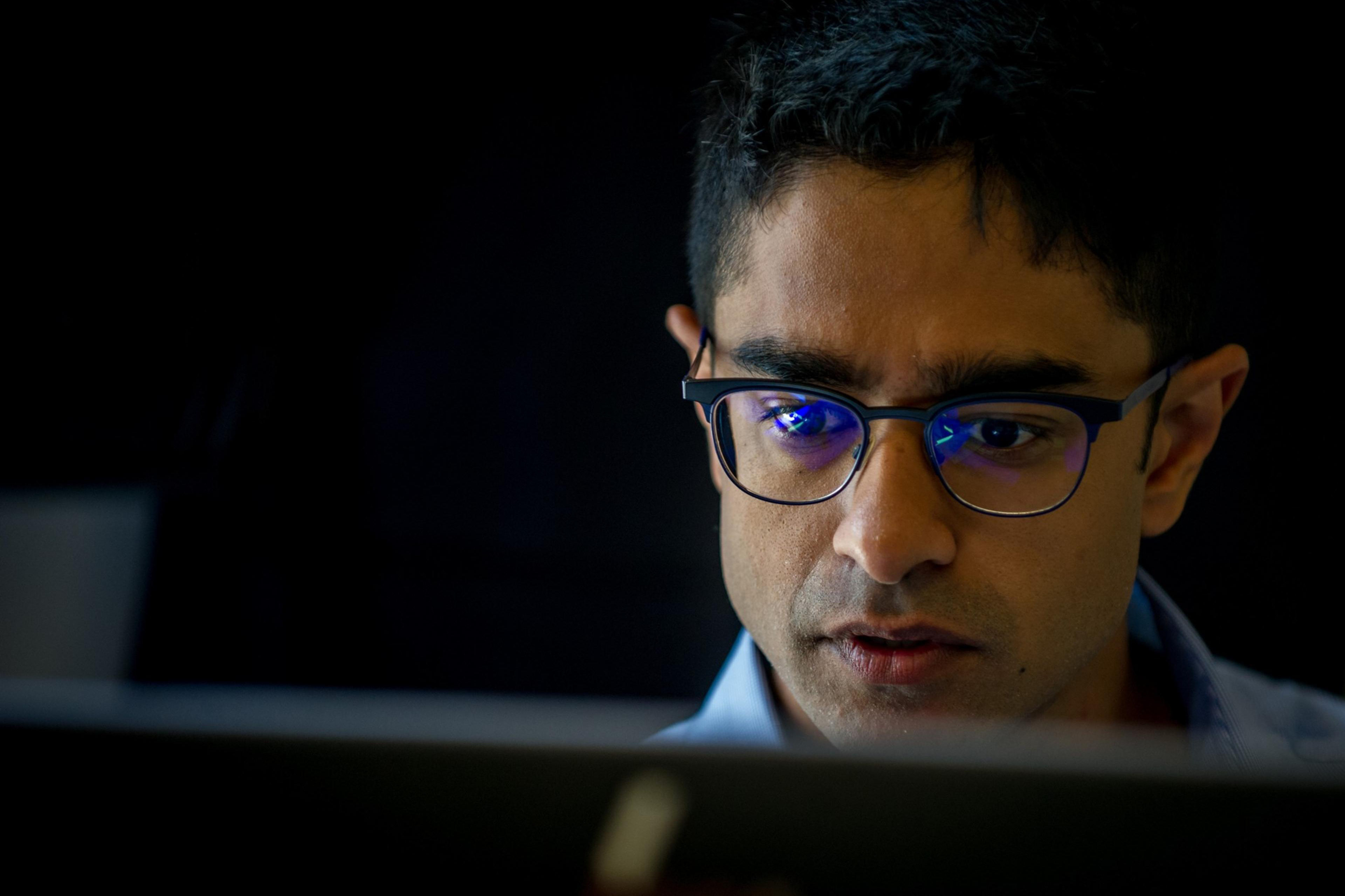 A person with glasses is intensely focused on something, likely a computer screen, which is reflected in the glasses. The background is dark and blurred.