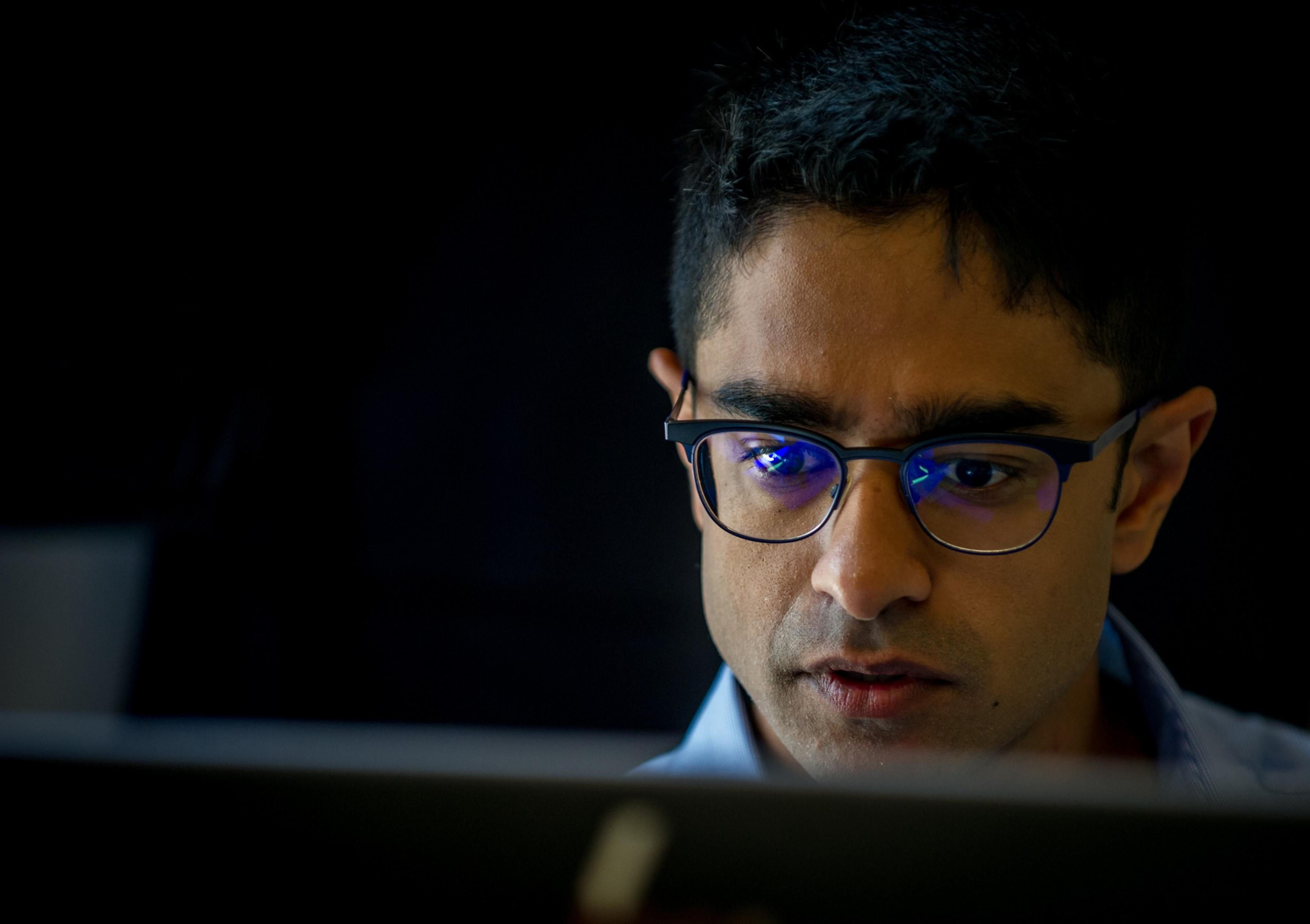 A person with glasses is intensely focused on something, likely a computer screen, which is reflected in the glasses. The background is dark and blurred.