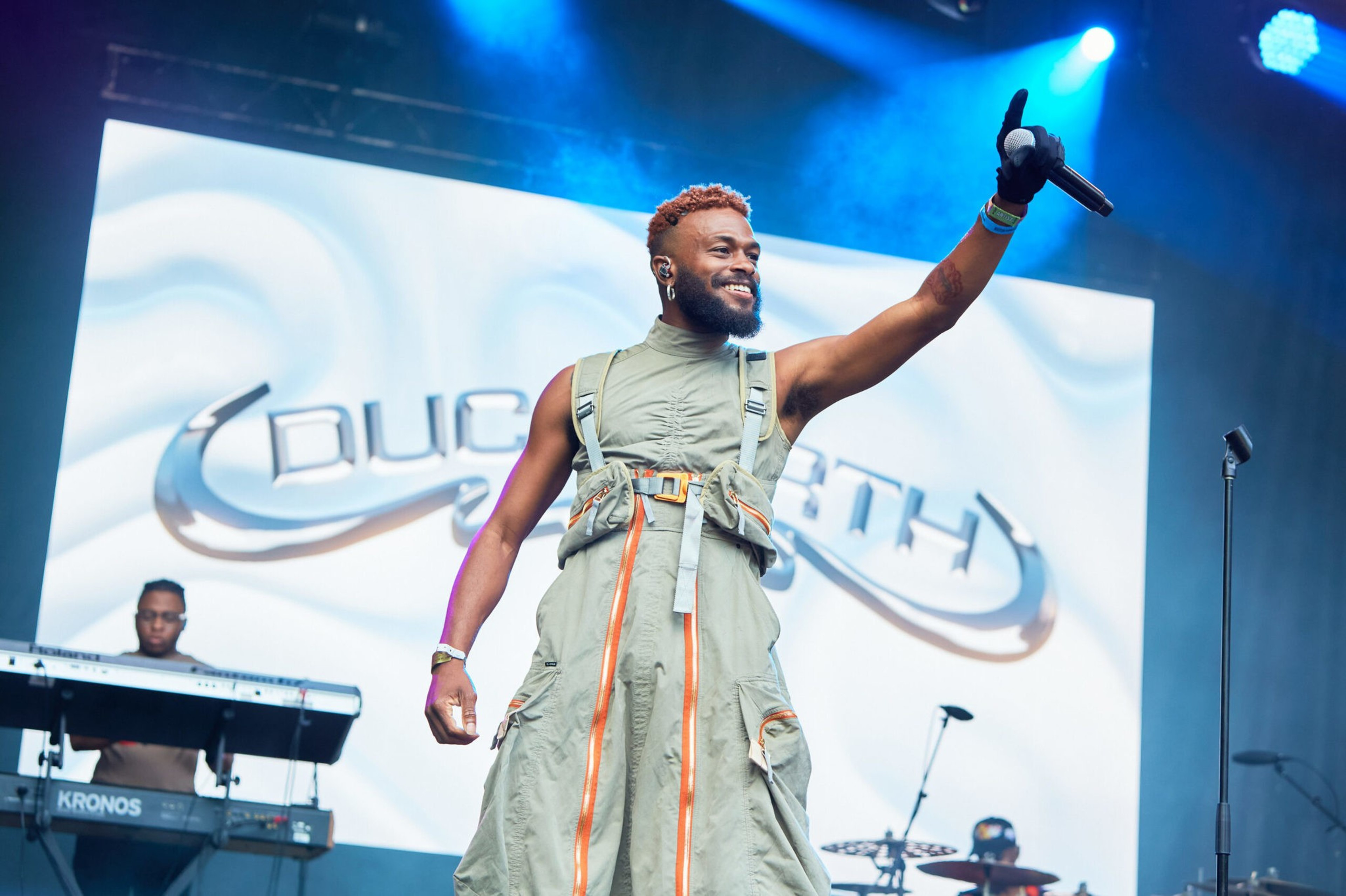 A smiling male performer raises a microphone with a gloved hand on stage, wearing a green outfit with orange accents, while a keyboard player performs in the background.