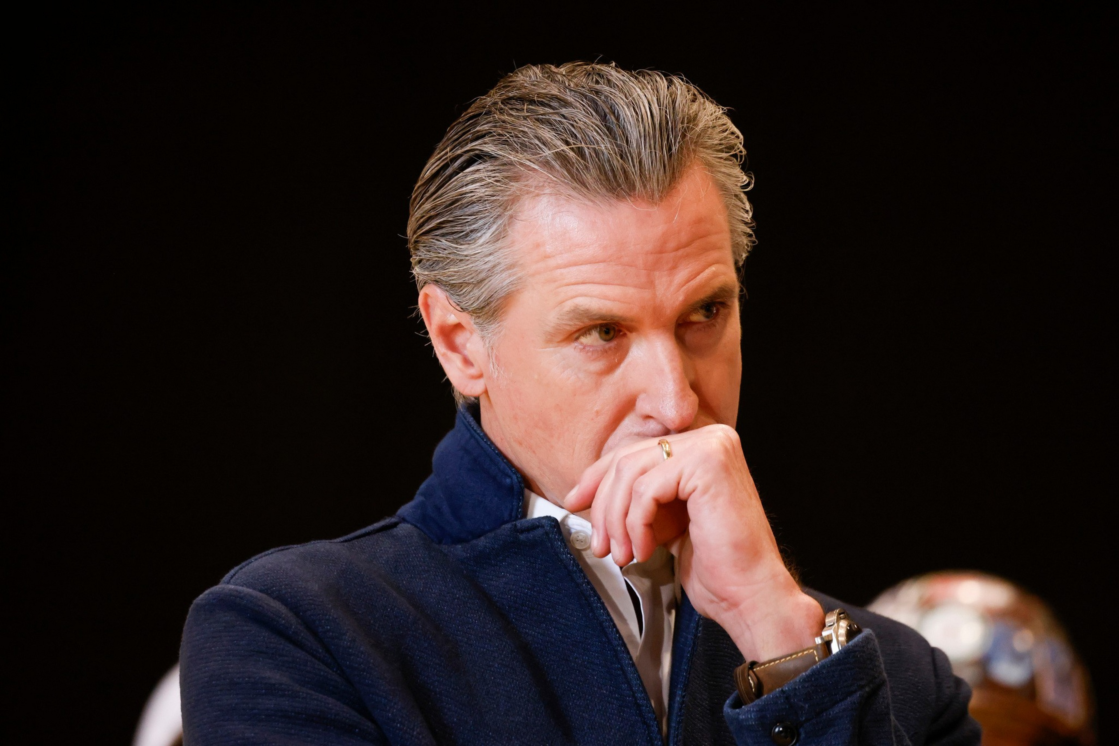 A middle-aged man with neatly combed gray hair wears a navy blue jacket and a watch, holding his hand near his mouth in a thoughtful pose.