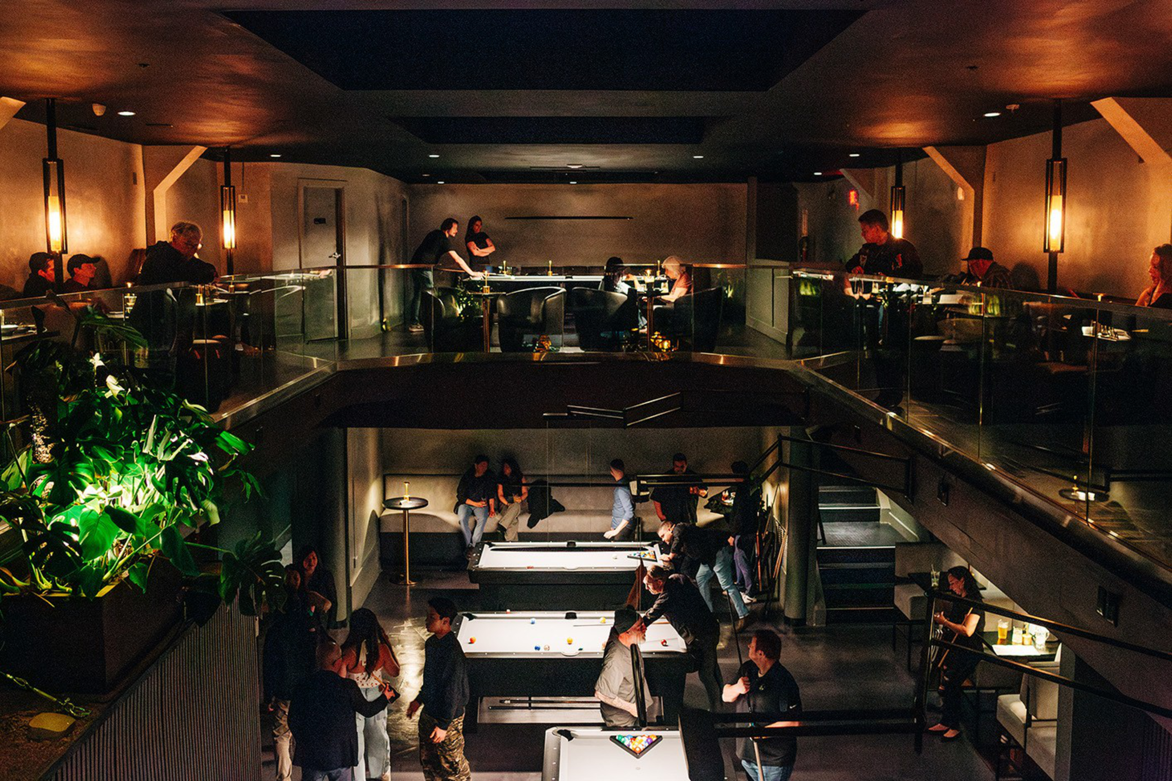 A dimly lit two-level lounge features people playing pool on the lower level and others seated or standing on the upper balcony with warm wall lights.