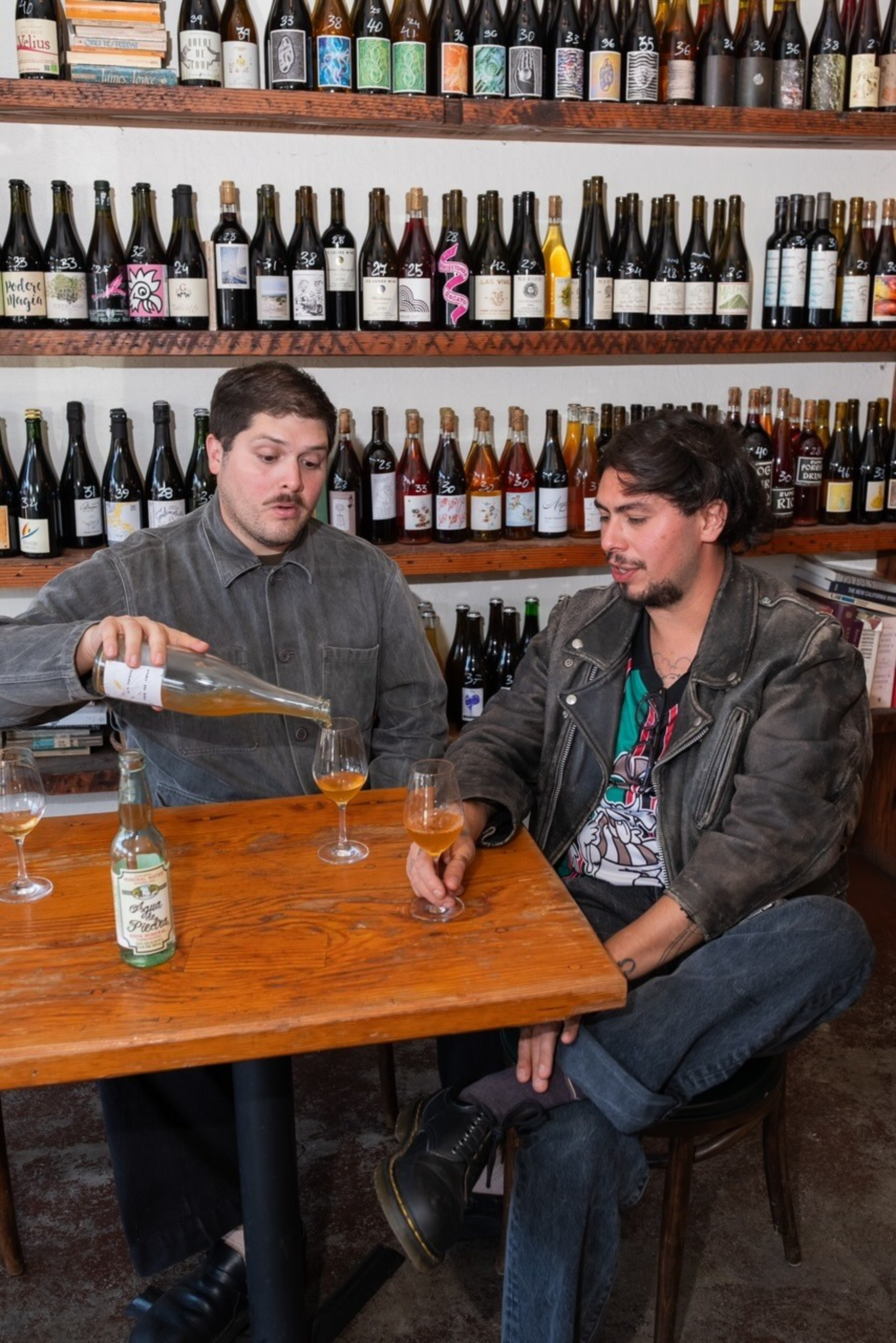 Two men sit at a wooden table in front of shelves filled with various wine bottles, one pouring a drink into a glass.