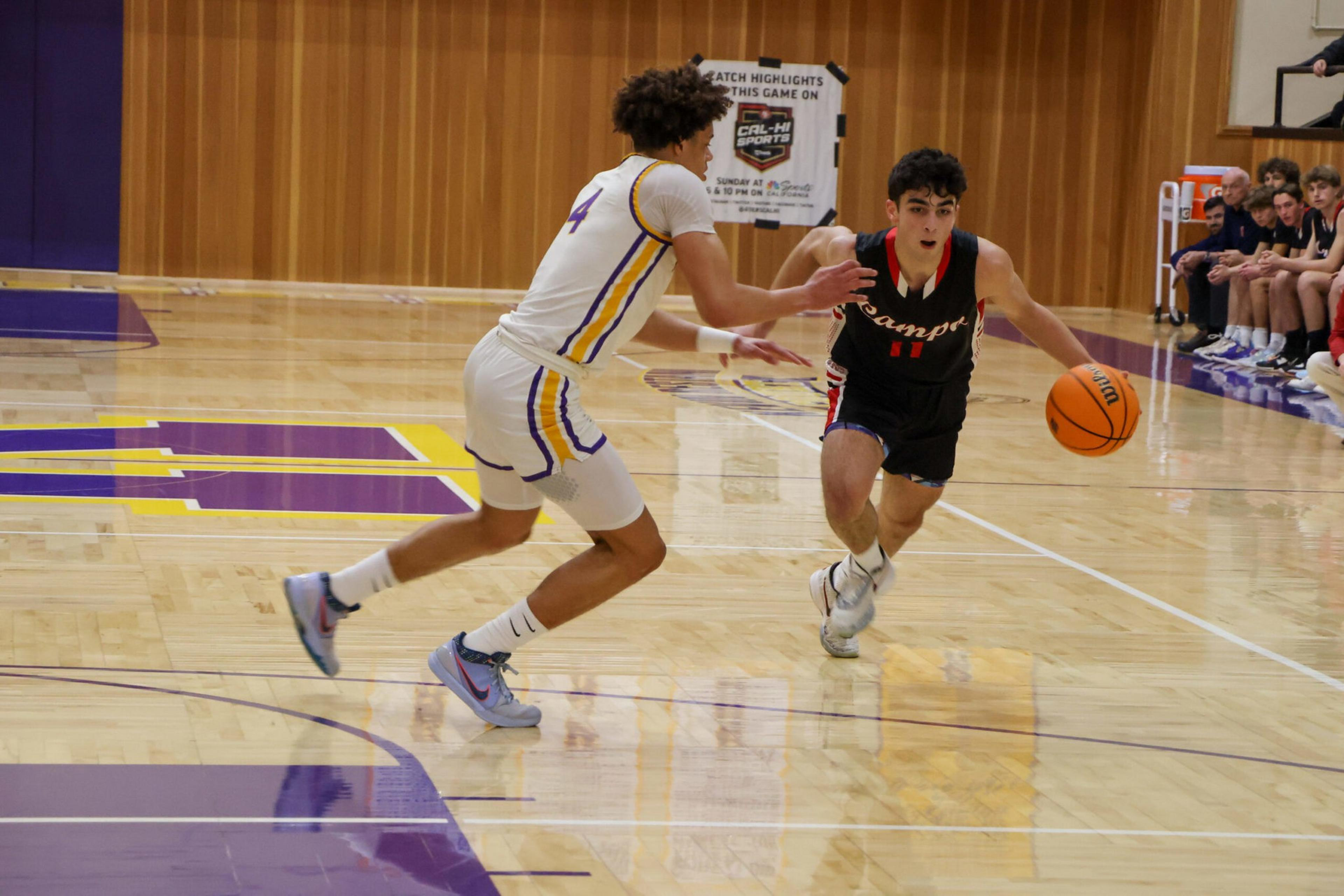 A basketball player in a black jersey dribbles past a defender in a white jersey on a polished indoor court with spectators seated on the side.