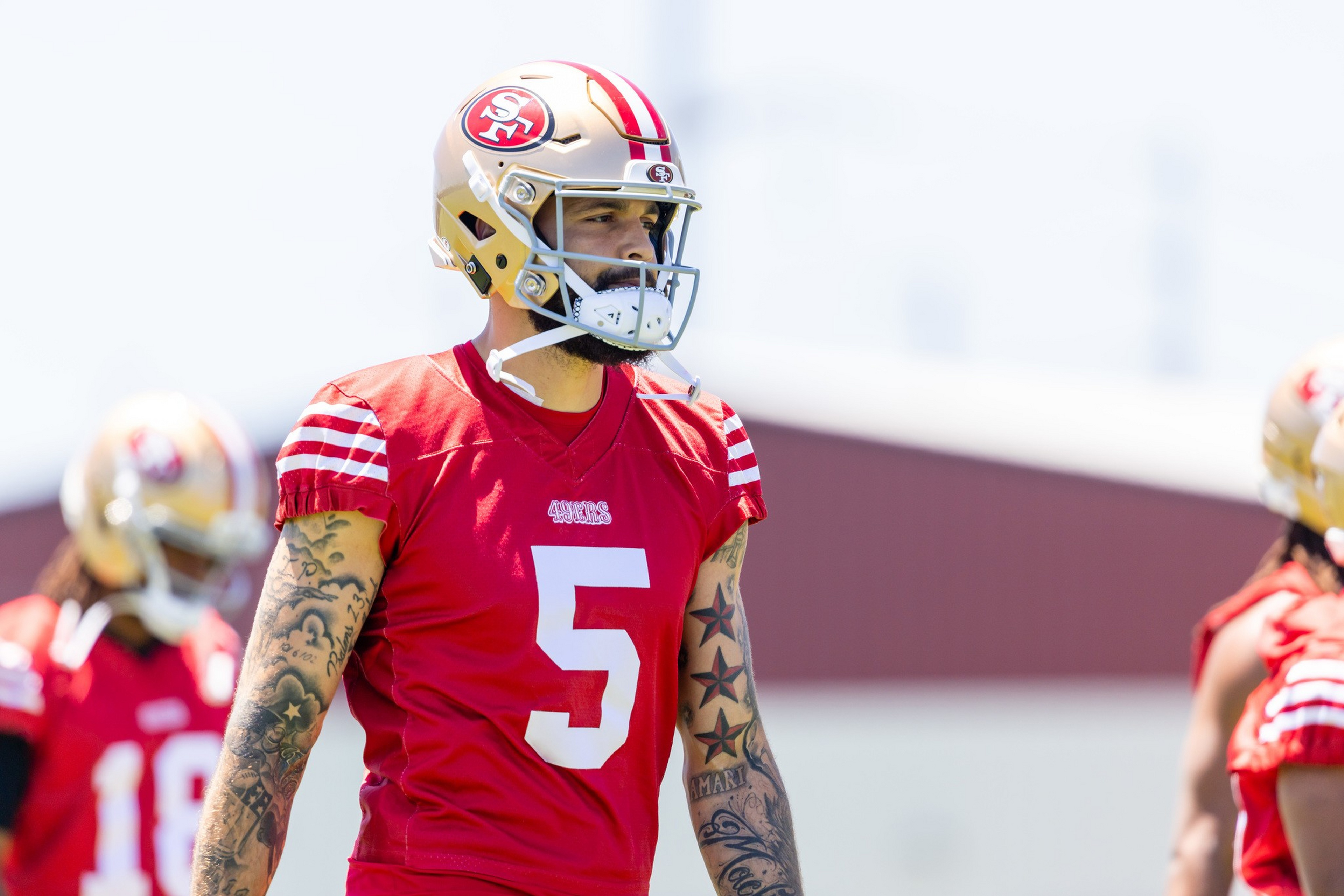 A football player wearing a red San Francisco 49ers jersey with number 5 and a gold helmet stands outdoors, showing multiple star tattoos on his arms.