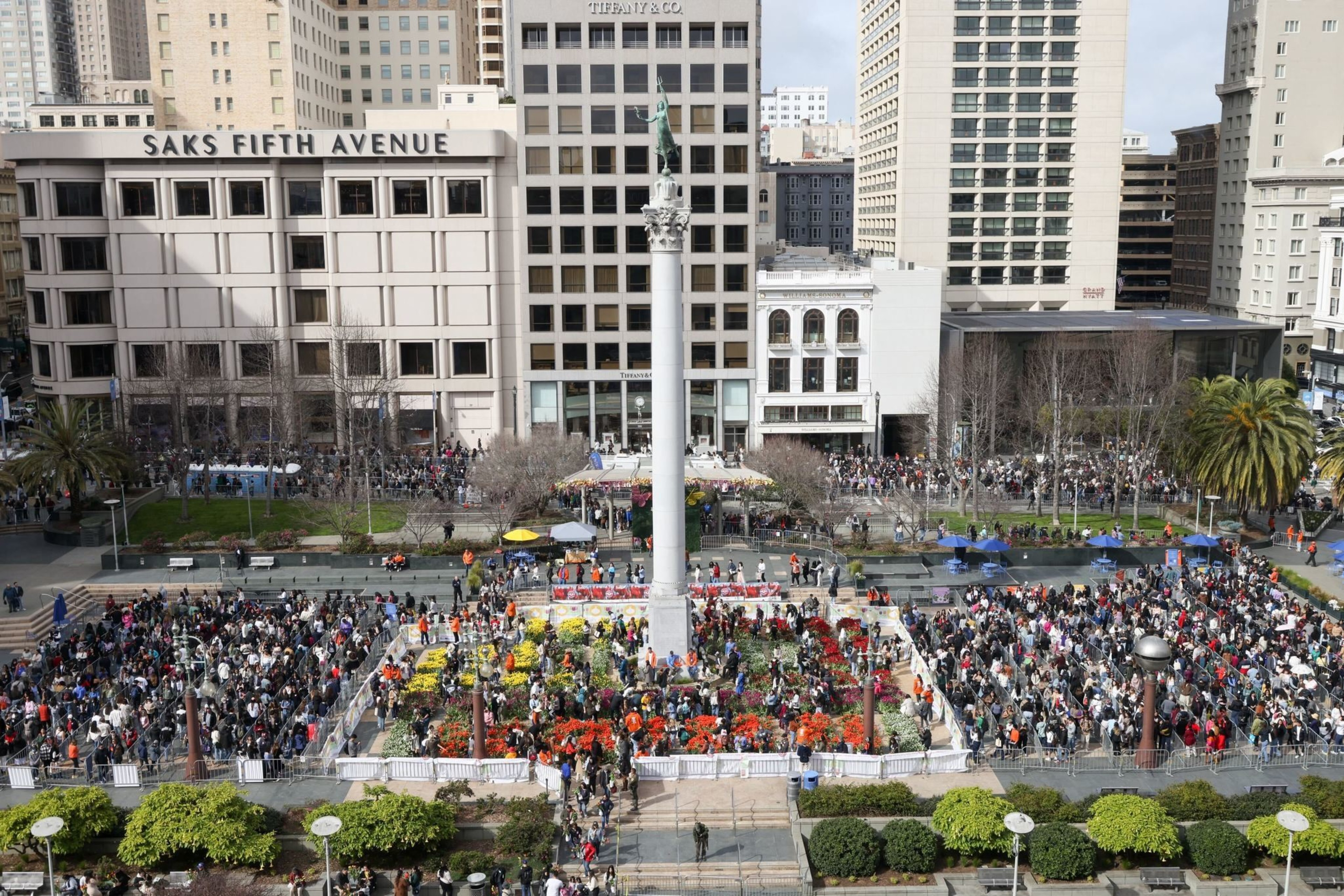 An aerial view of a busy city square with a crowd, colorful flowers, and surrounding buildings.