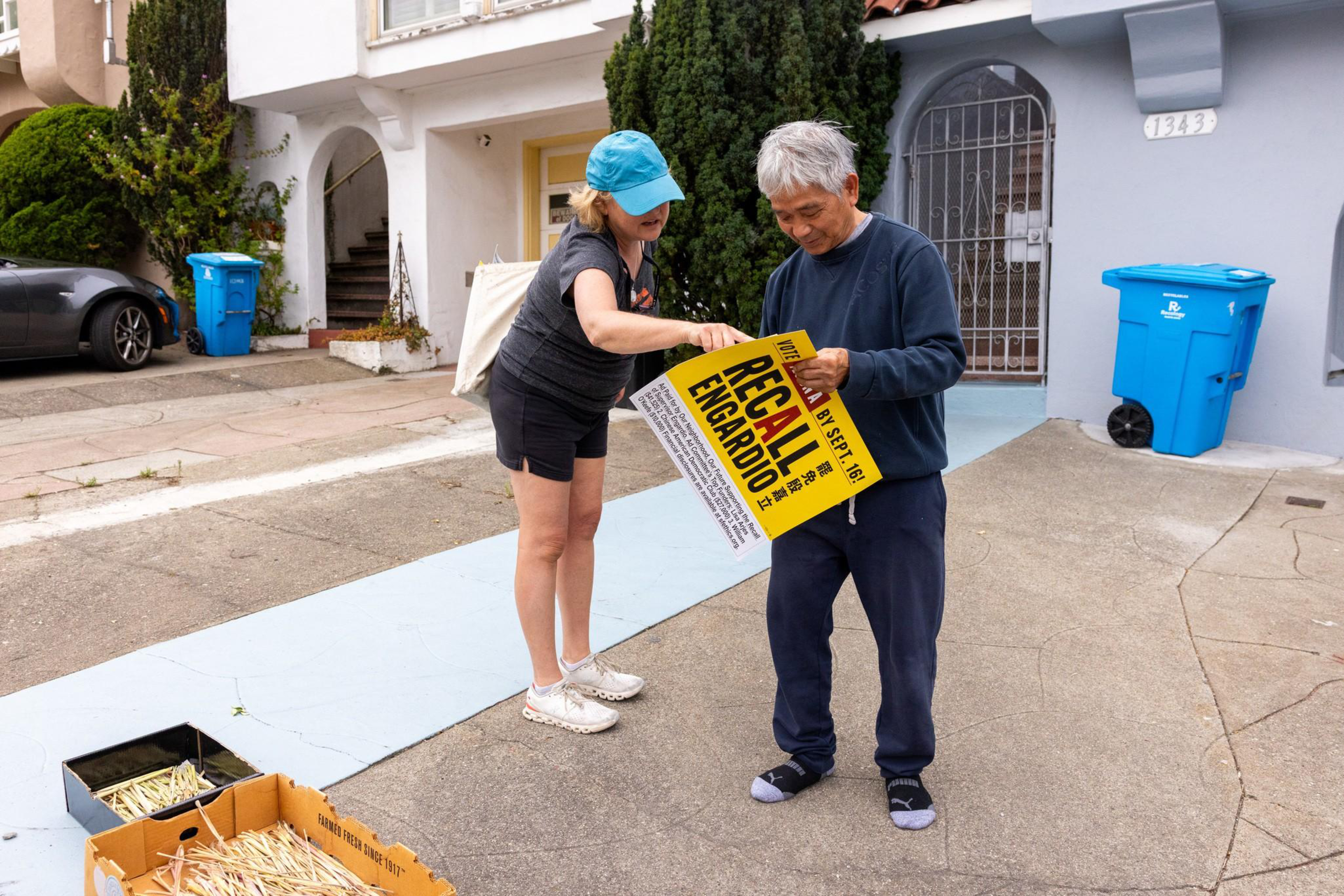 A young person wearing a blue hairnet hands a “Recall Newsom” sign to an older person outside a house on a residential street.