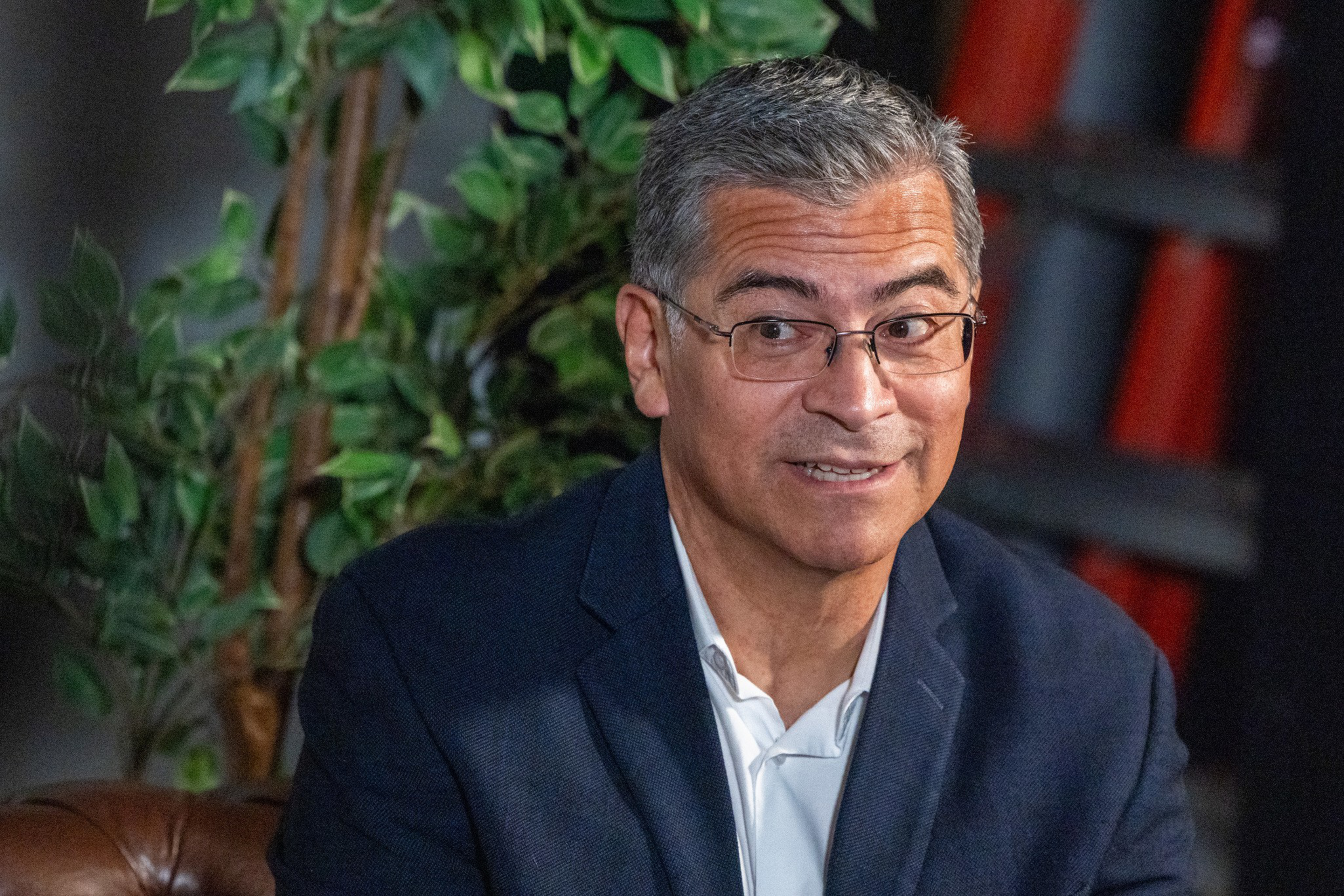 A man with short gray hair and glasses wears a dark blazer over a white shirt, sitting and smiling slightly with a leafy plant behind him.