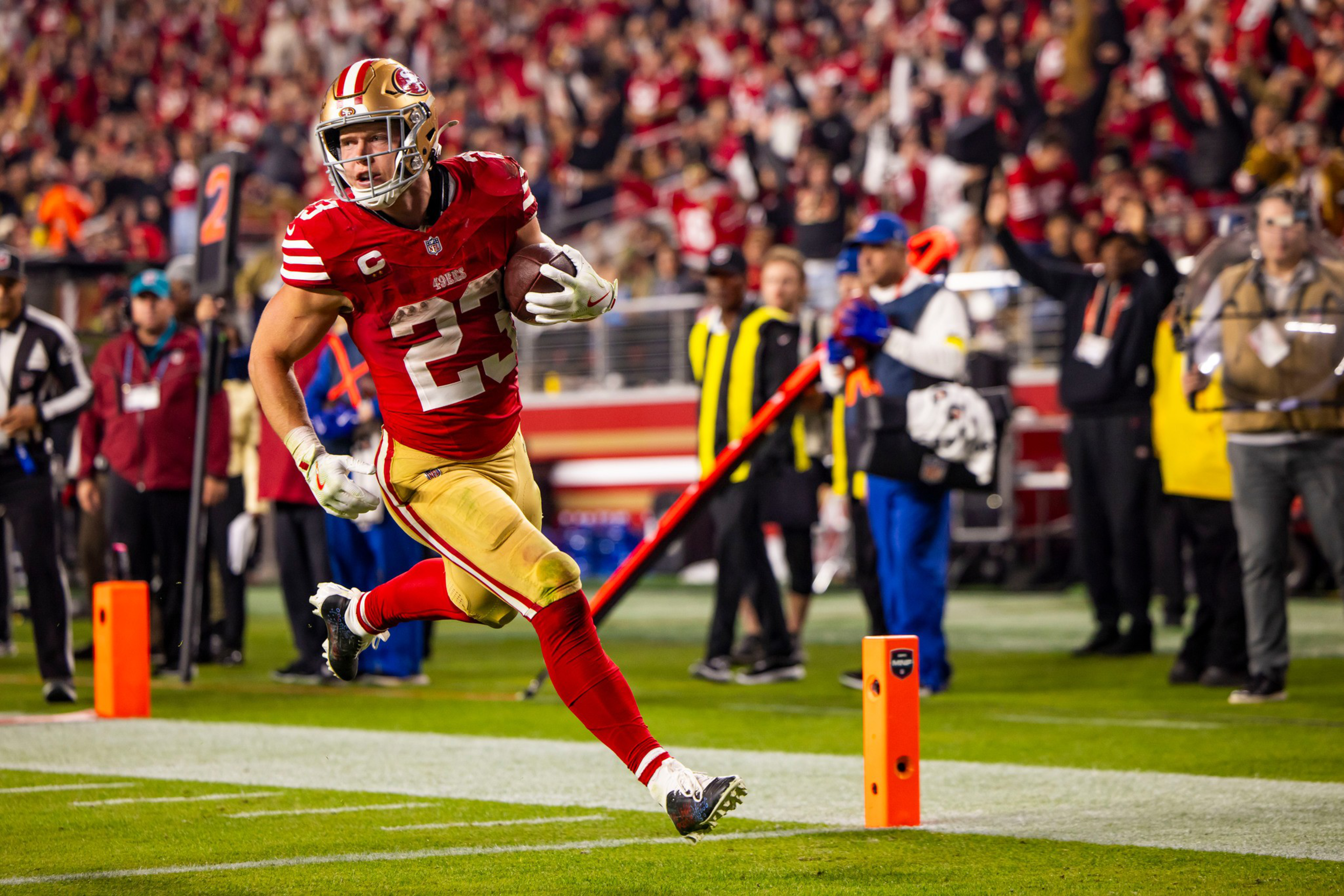 A football player in a red and gold uniform, number 23, runs with the ball toward the end zone during a game, with a crowd and officials in the background.