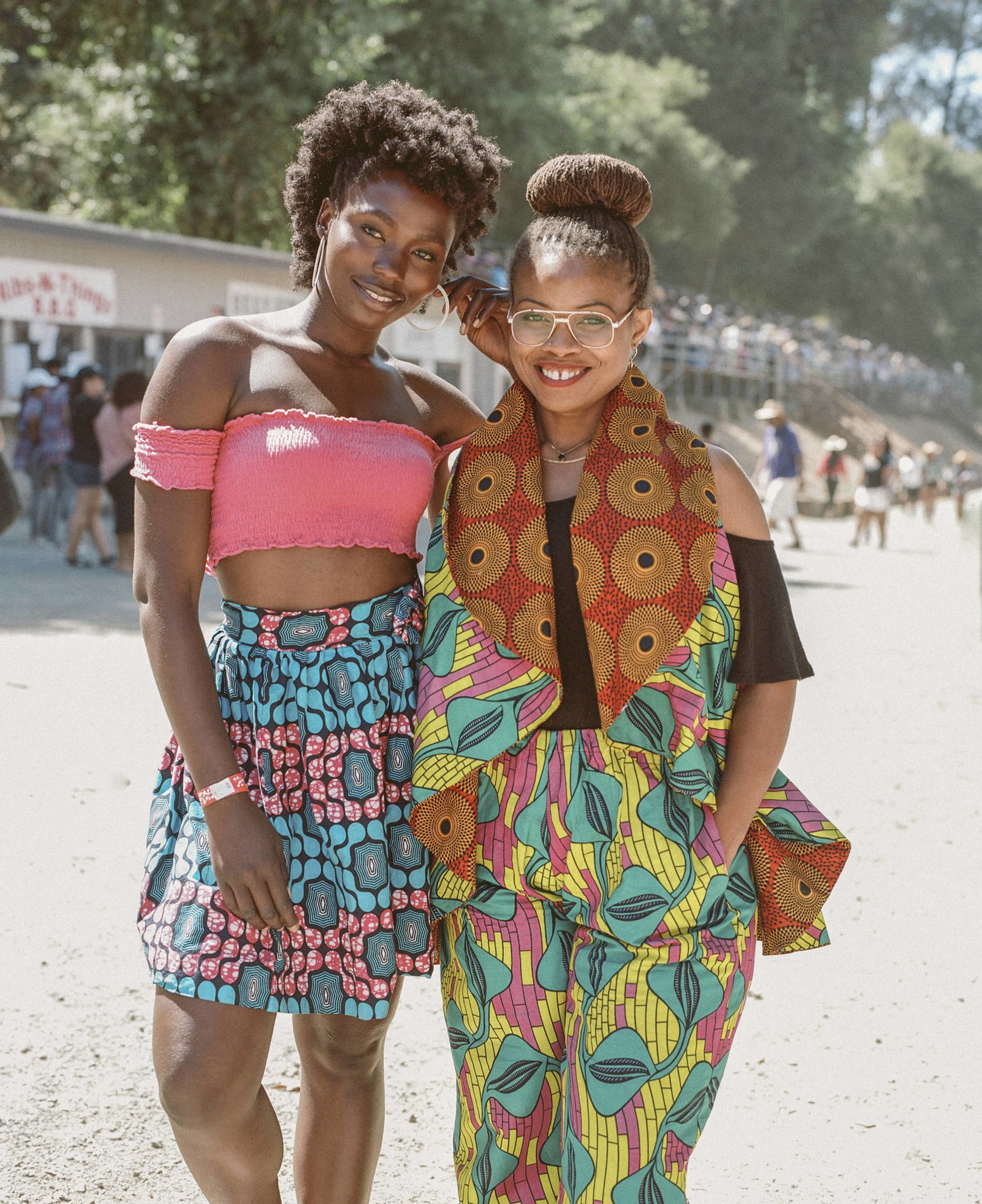 Two women are smiling outdoors. One wears a pink top with a blue patterned skirt; the other wears colorful, patterned attire with glasses. Trees are in the background.