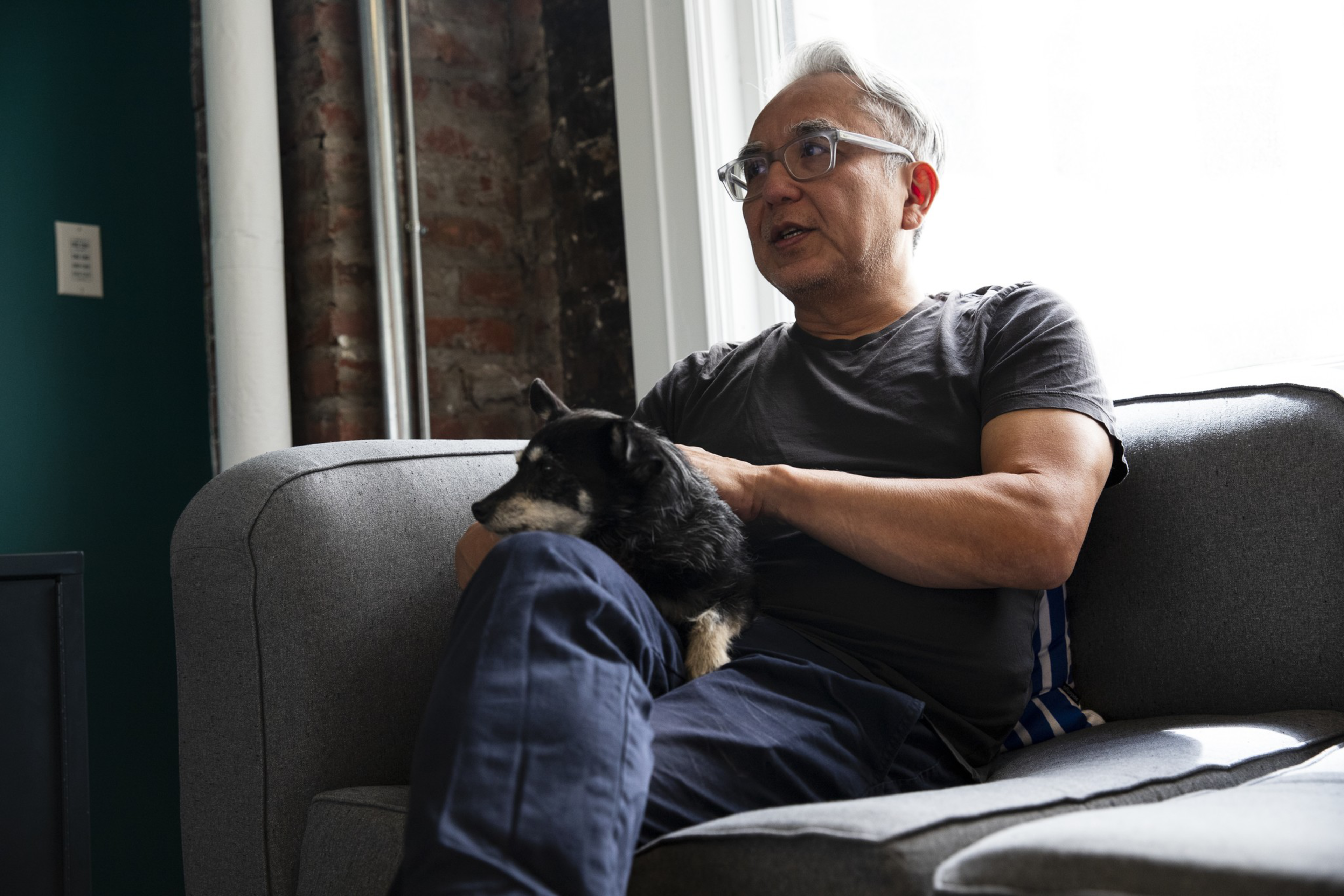 An older man wearing glasses and a dark shirt sits on a gray couch, holding a small black dog on his lap near a window.