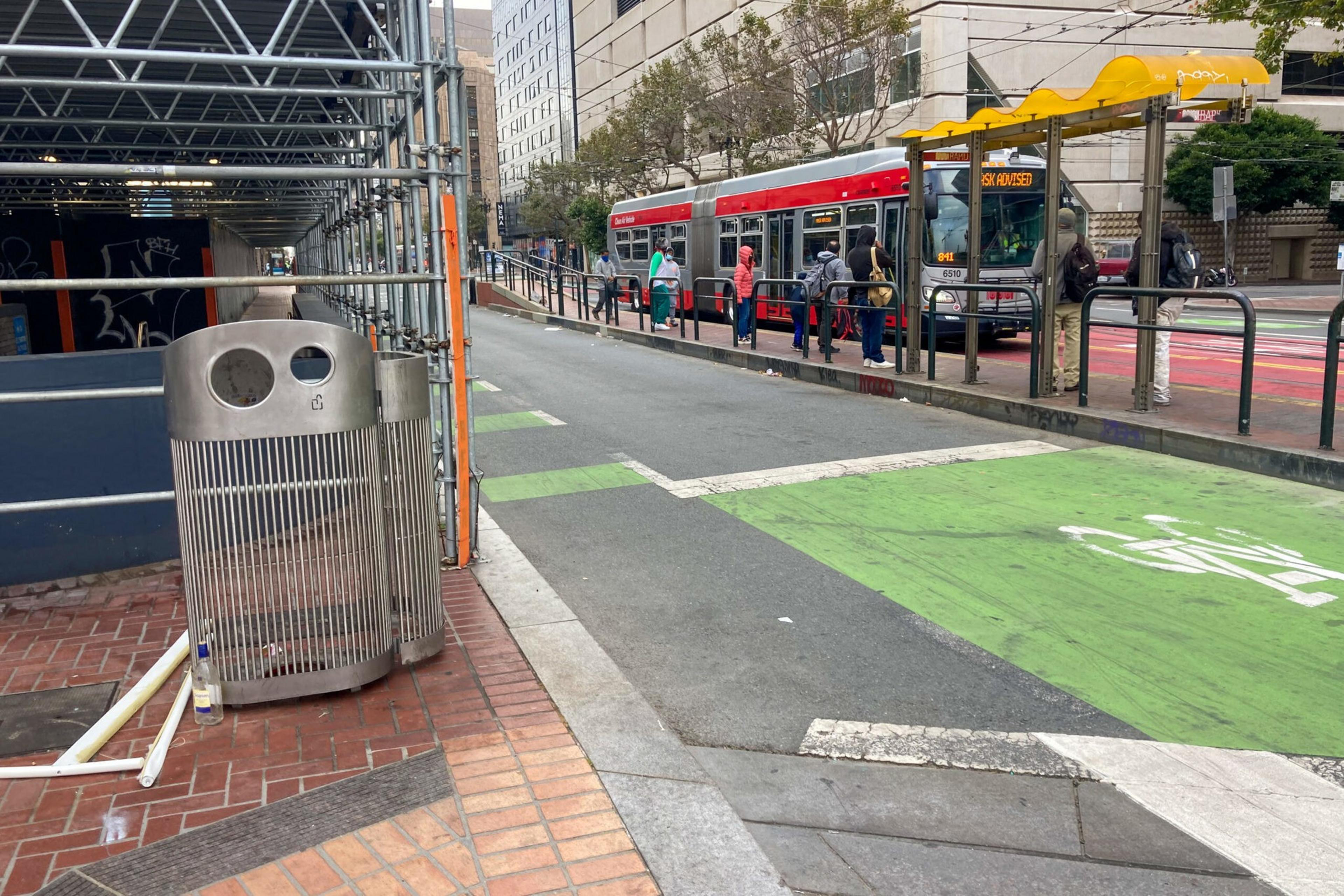 A street with a red tram at a stop, passengers waiting on the platform, and a trash can next to scaffolding on the sidewalk. The road has green-marked bike lanes.