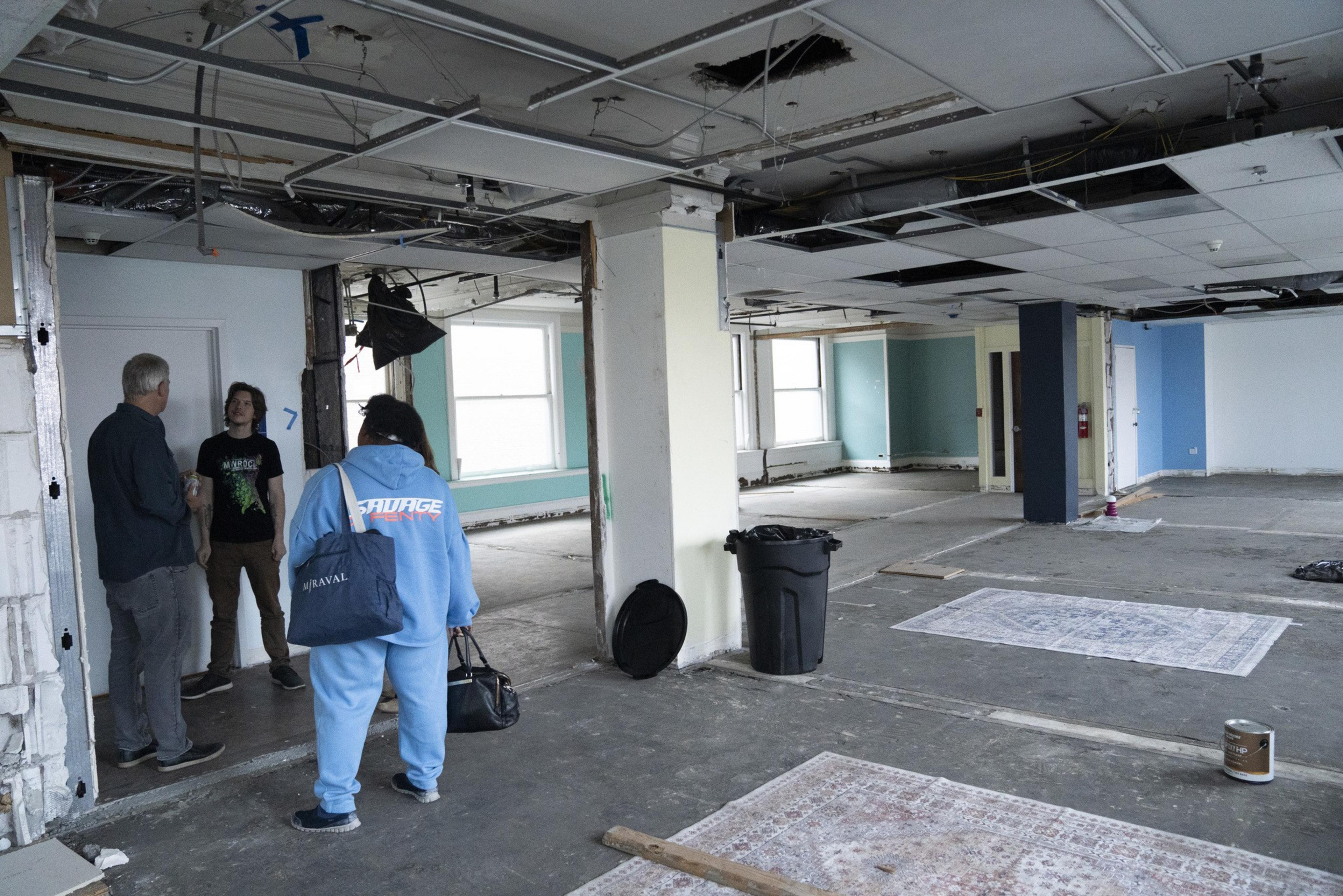 Three people stand in a partially demolished room with exposed ceilings and unfinished floors. Rugs and a trash can are on the floor, with walls painted teal and white.