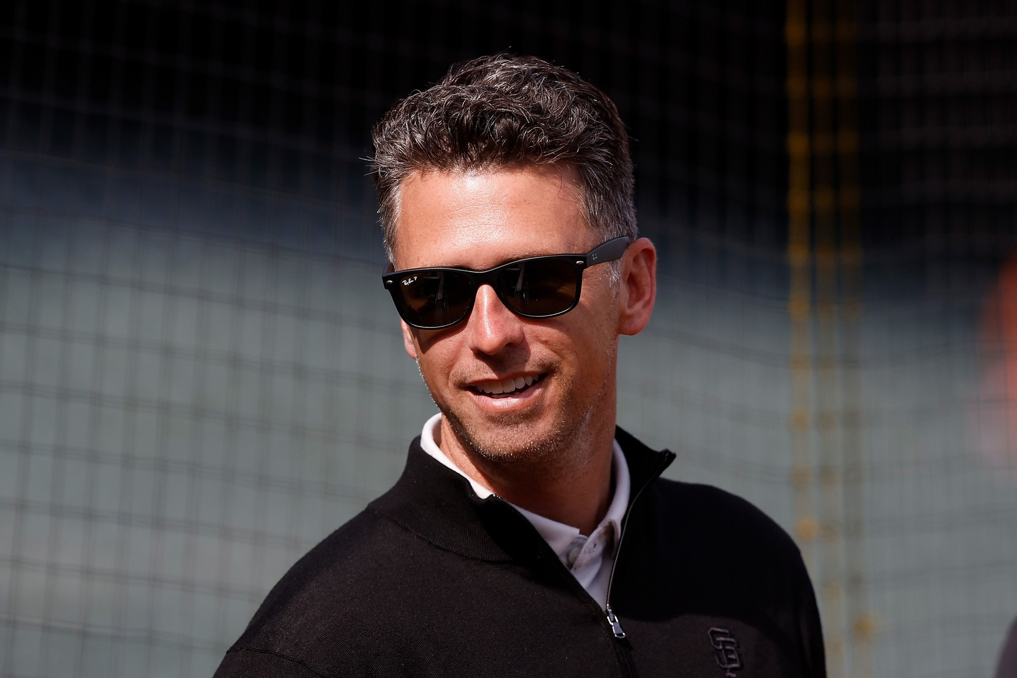 A man with short, curly hair wearing black sunglasses and a black zip-up jacket smiles slightly against a blurred netted background.