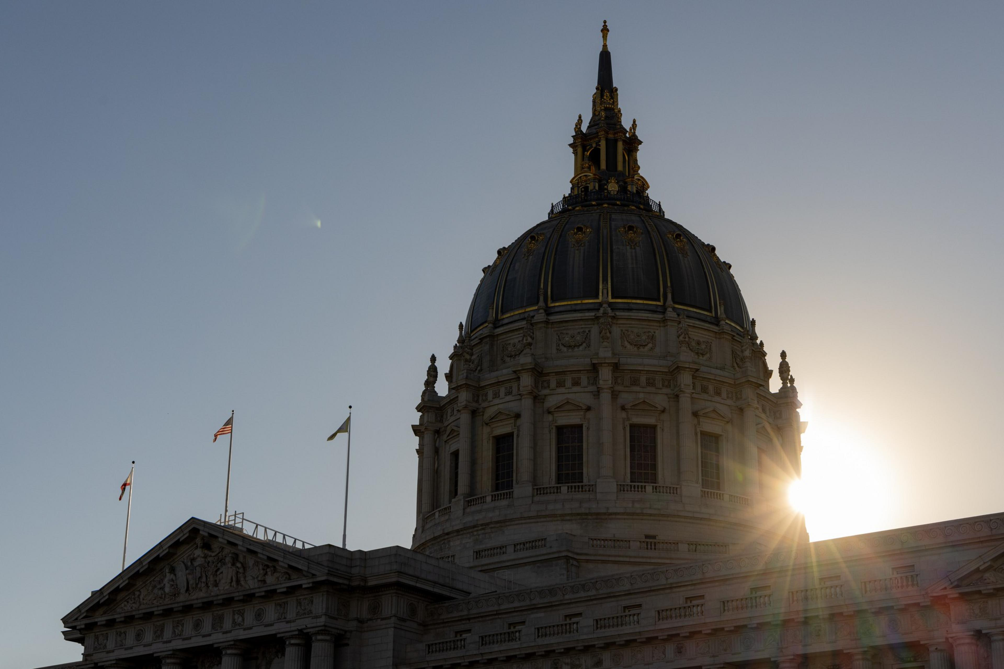 The image shows the dome of a grand building silhouetted against the setting sun, with two flags and clear skies in the background.