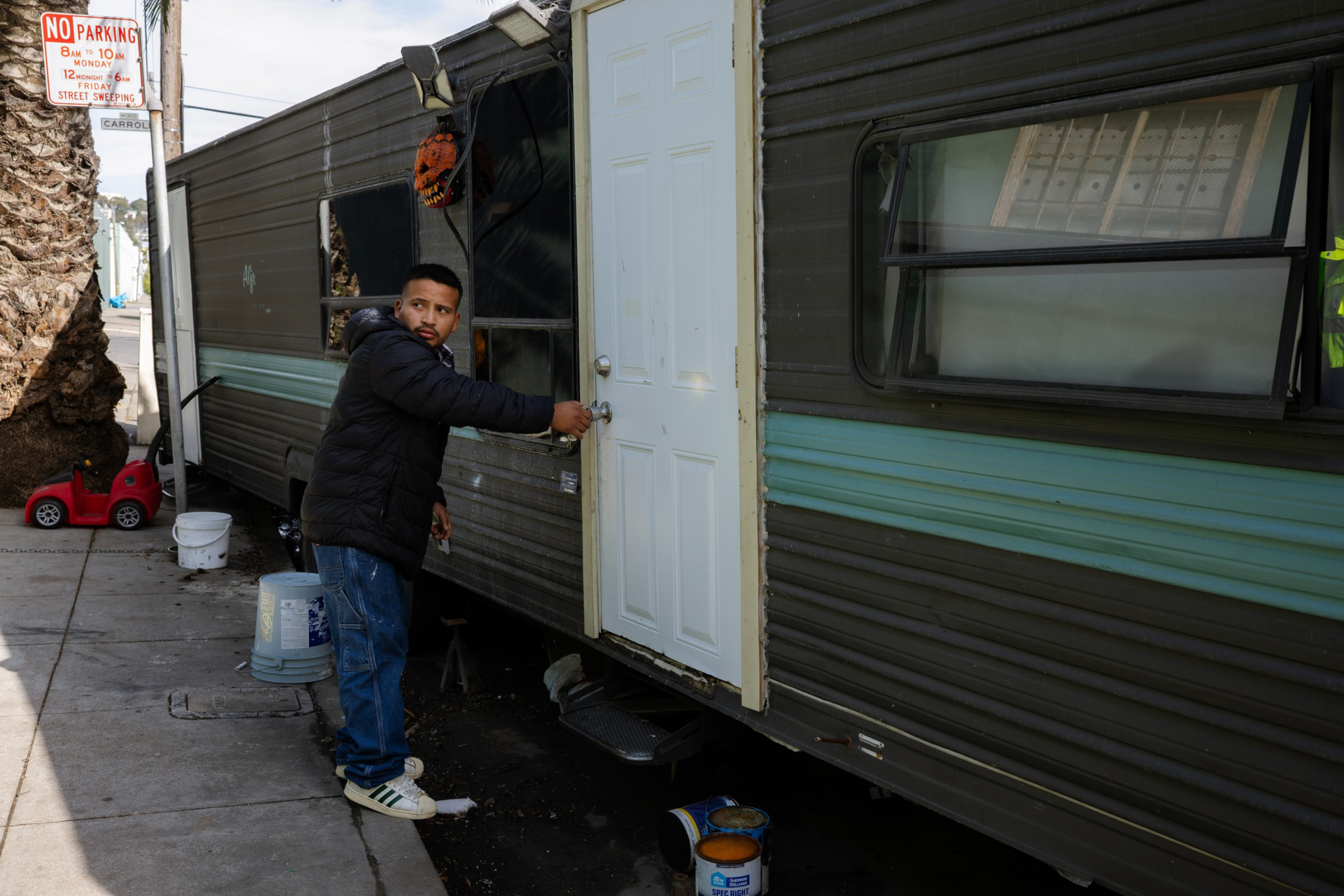 A man in a black jacket and jeans is unlocking a white door on a dark brown trailer with a blue stripe, parked on a sidewalk near a palm tree.