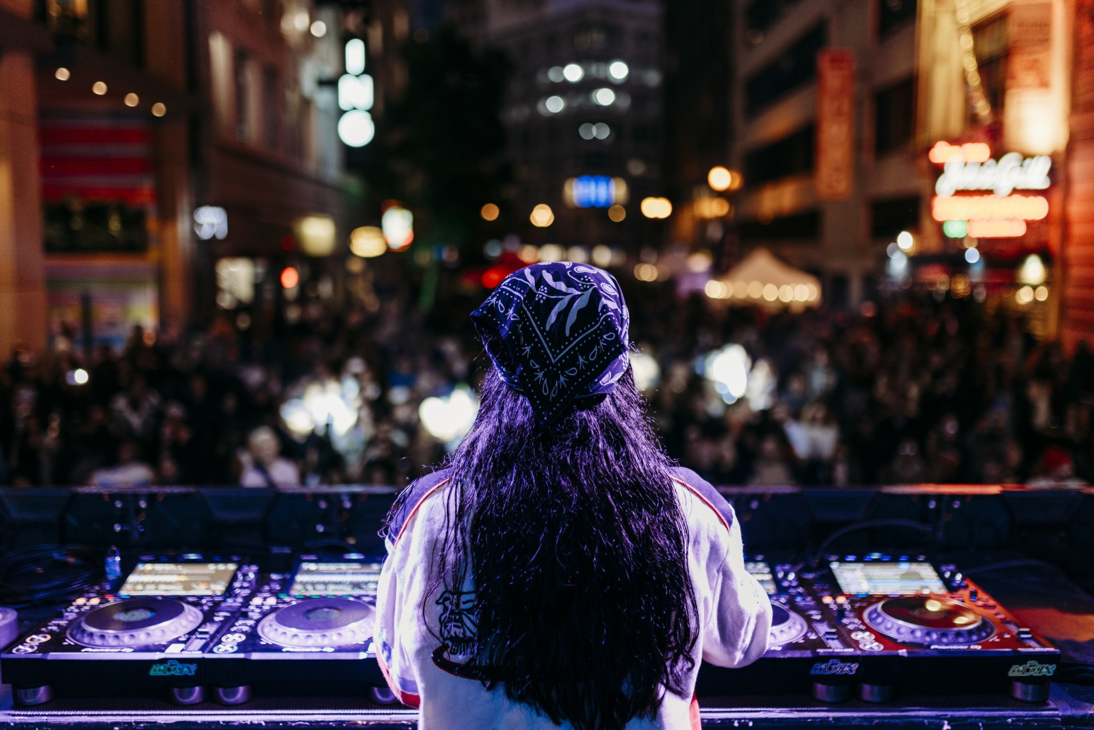 A DJ with long hair and a bandana performs at night in front of a large, blurred crowd in a city street filled with bright lights and buildings.